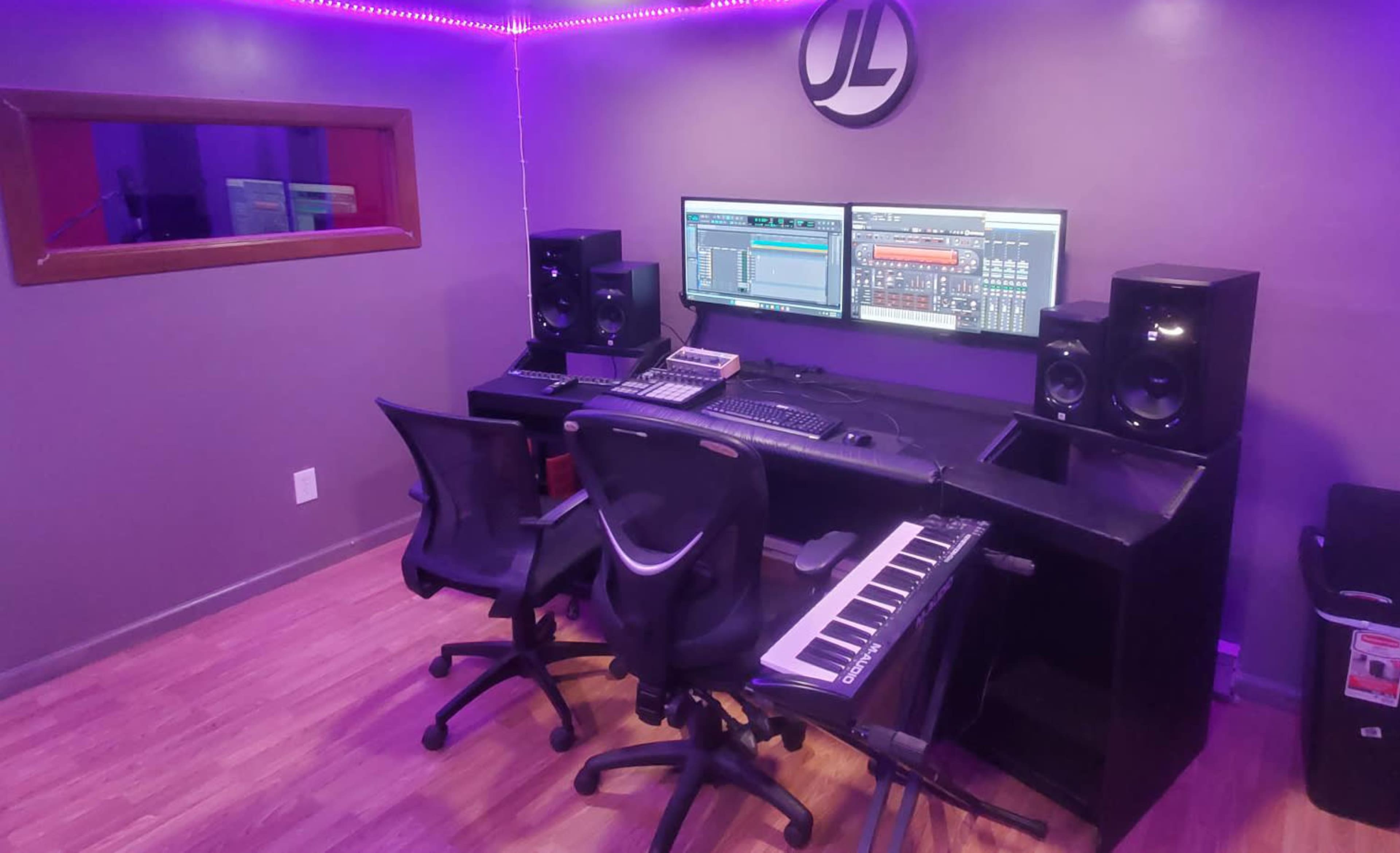 A modern music production studio featuring a dual monitor setup, audio speakers, a keyboard, and two rolling chairs.