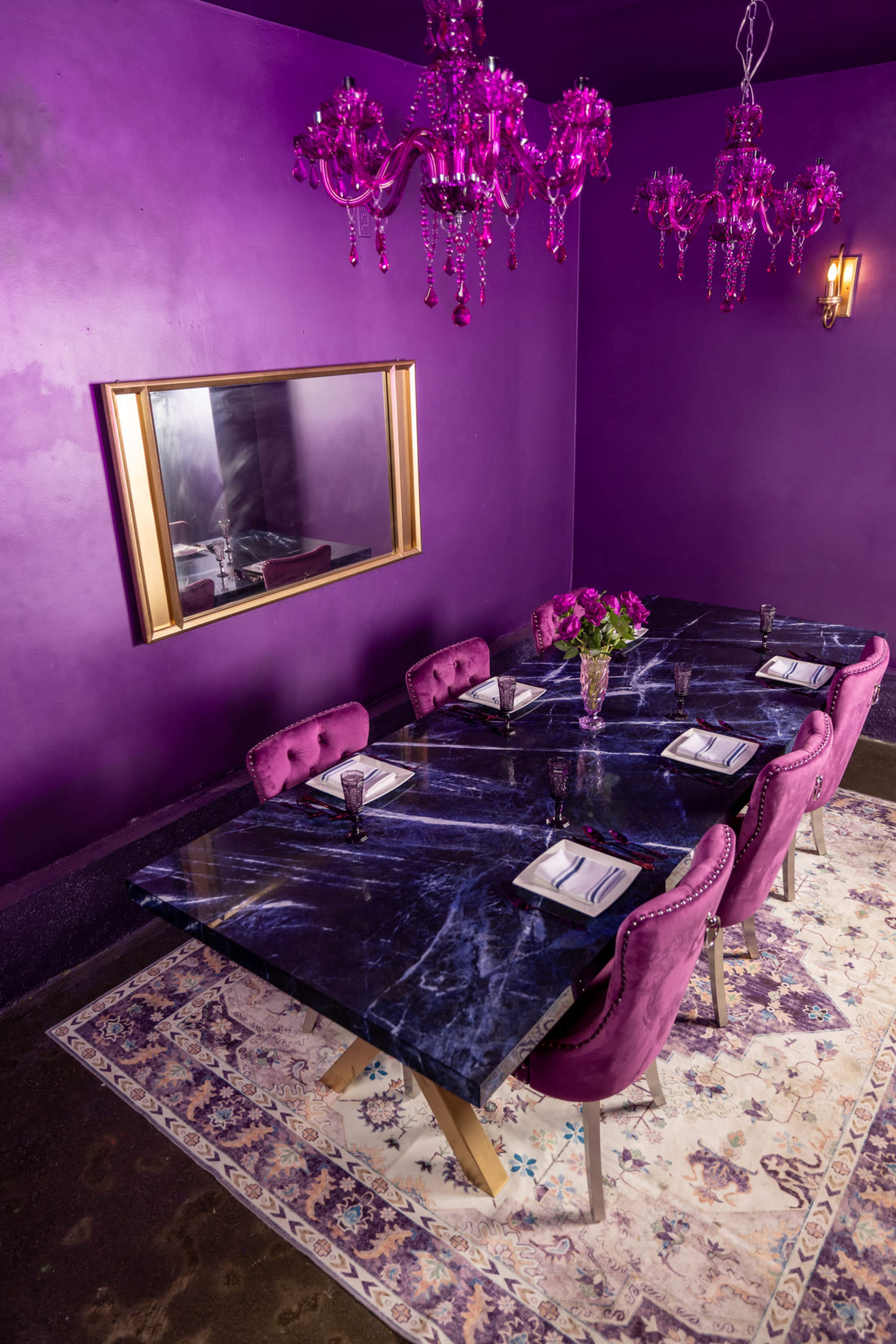 A large marble dining table with eight purple velvet chairs is set on a decorative rug in a room with purple walls and adorned with pink chandeliers.