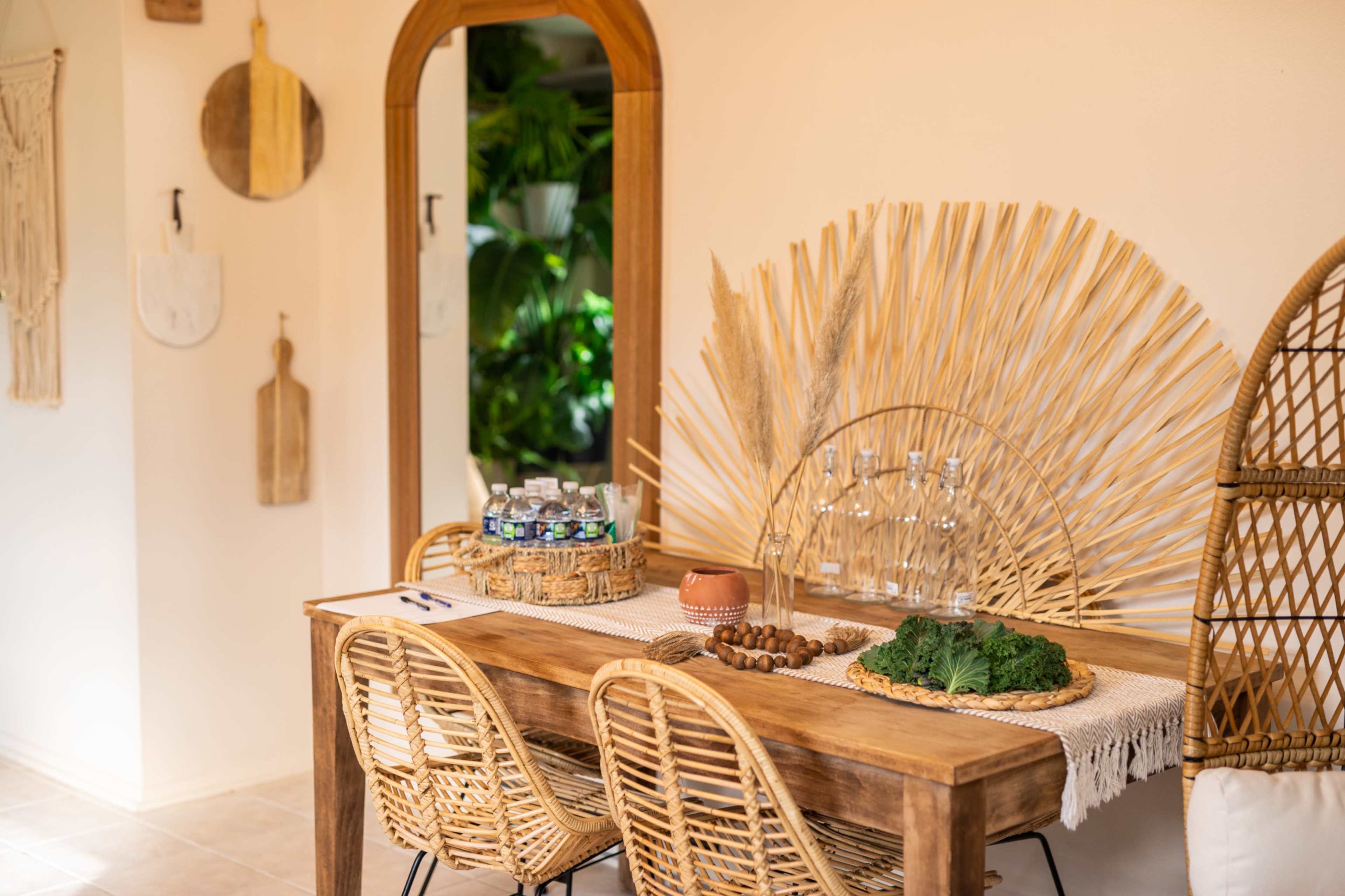A rustic wooden dining table is set with glass bottles, a bowl of kale, and decorative elements, surrounded by wicker chairs in a warmly lit room.