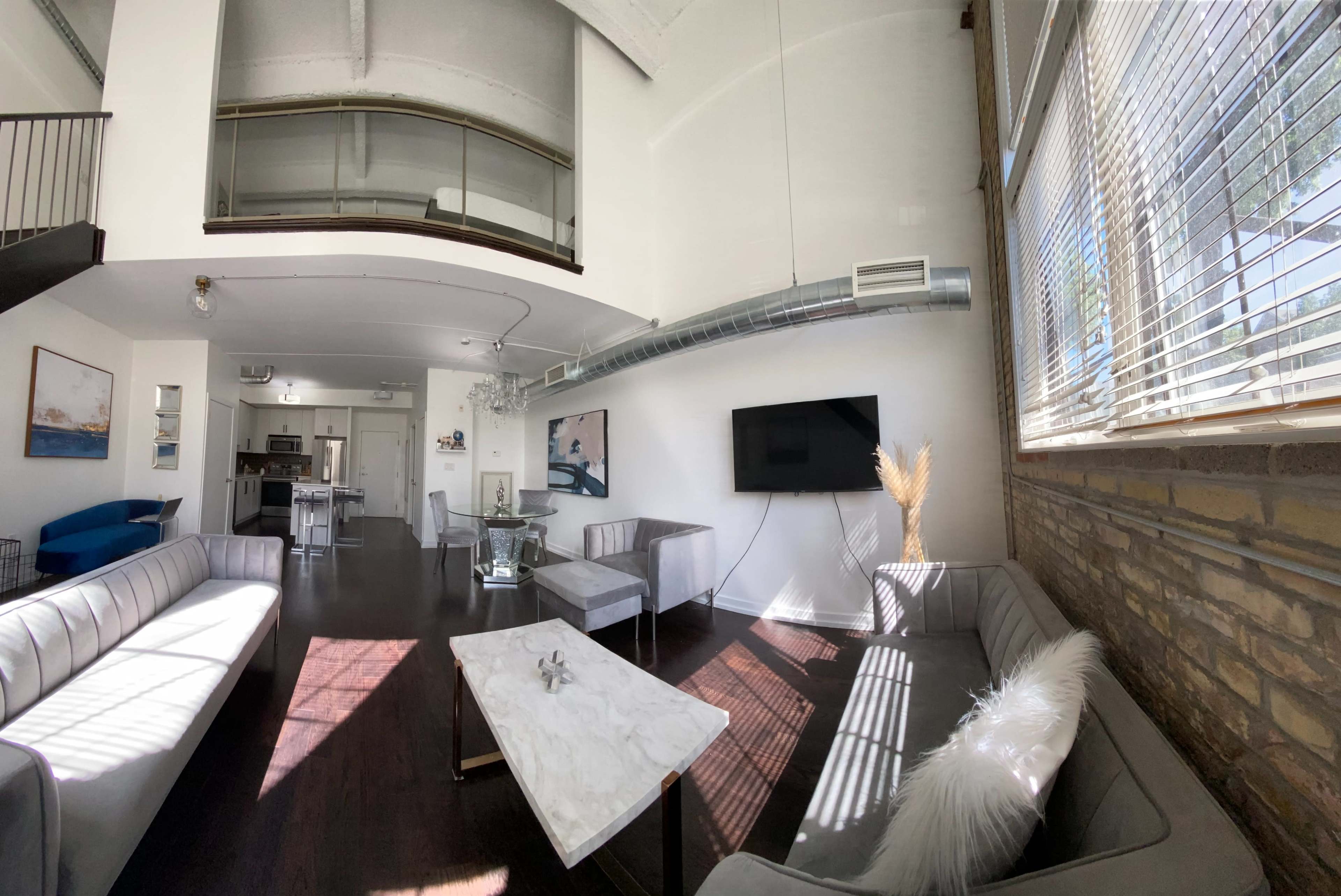 A spacious, modern living area with high ceilings, featuring gray sofas, a coffee table, and a large television mounted on the wall, with a kitchen visible in the background.