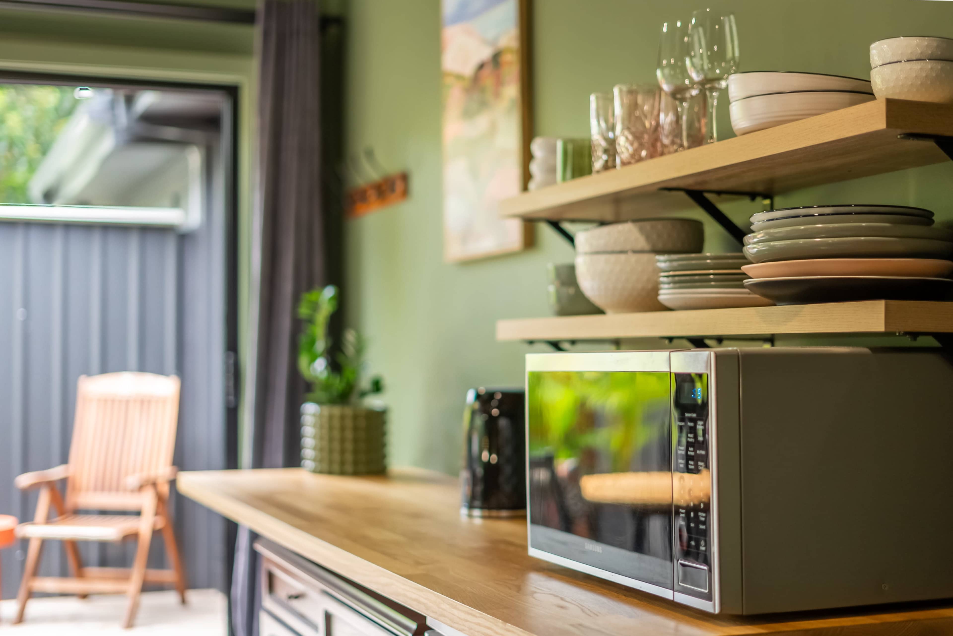 A kitchen countertop features a microwave, stacked plates, glasses, and a potted plant against a green wall.