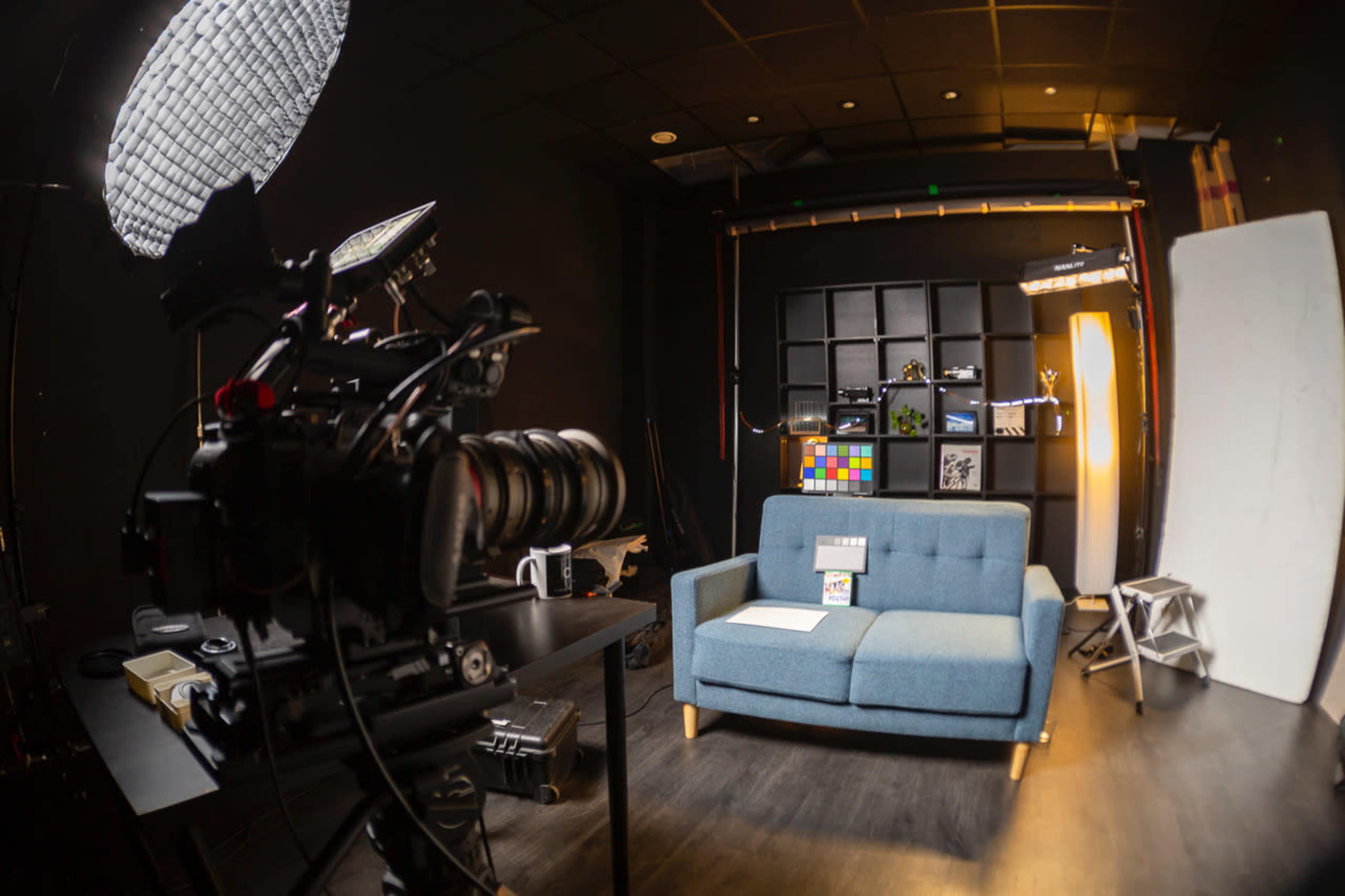 A camera is positioned in front of a blue couch set against a black wall with shelves, studio lights, and an array of colorful objects displayed.