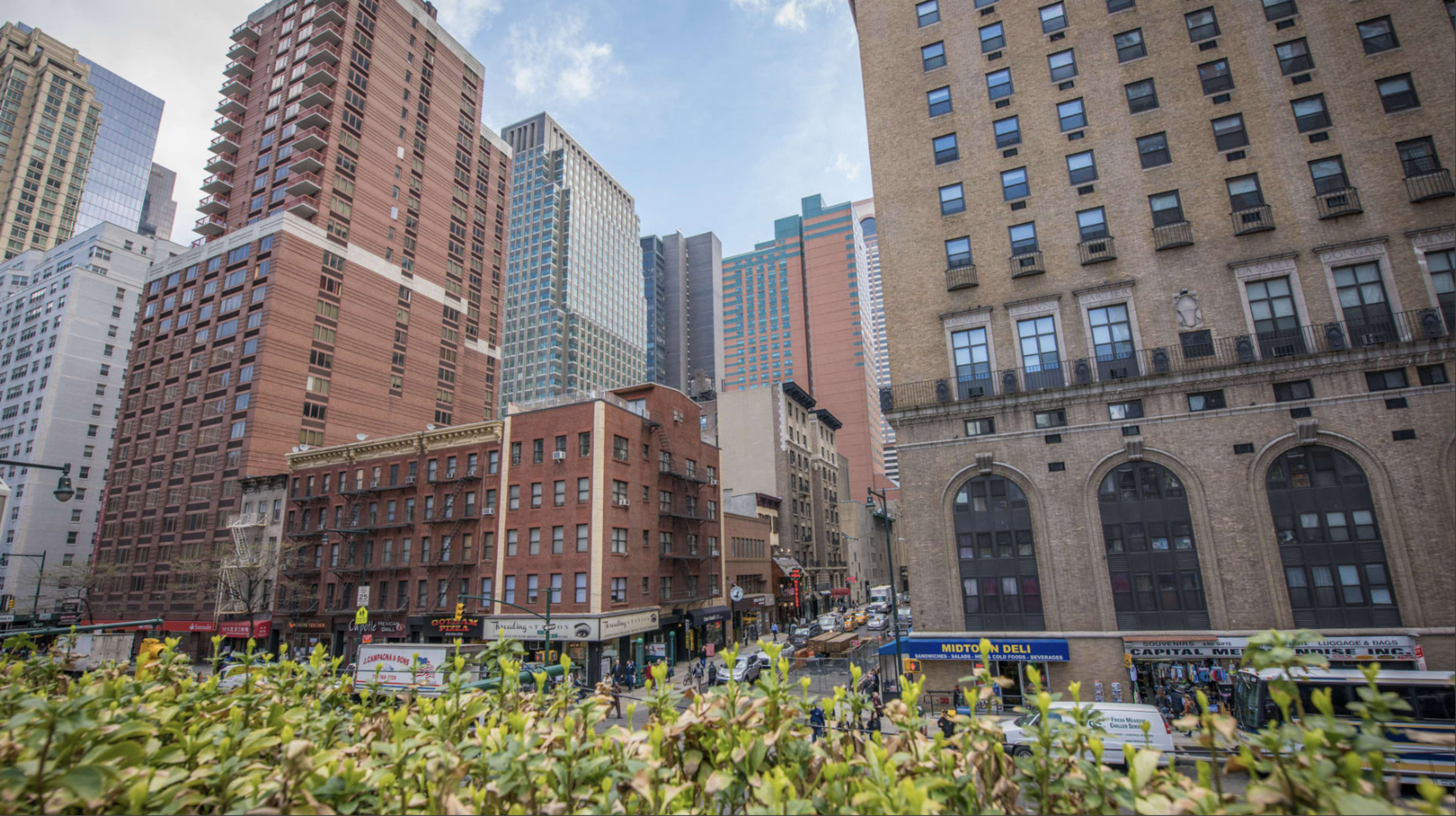 The image shows a bustling city street in New York, lined with a mix of historic and modern buildings, filled with pedestrians and vehicles.