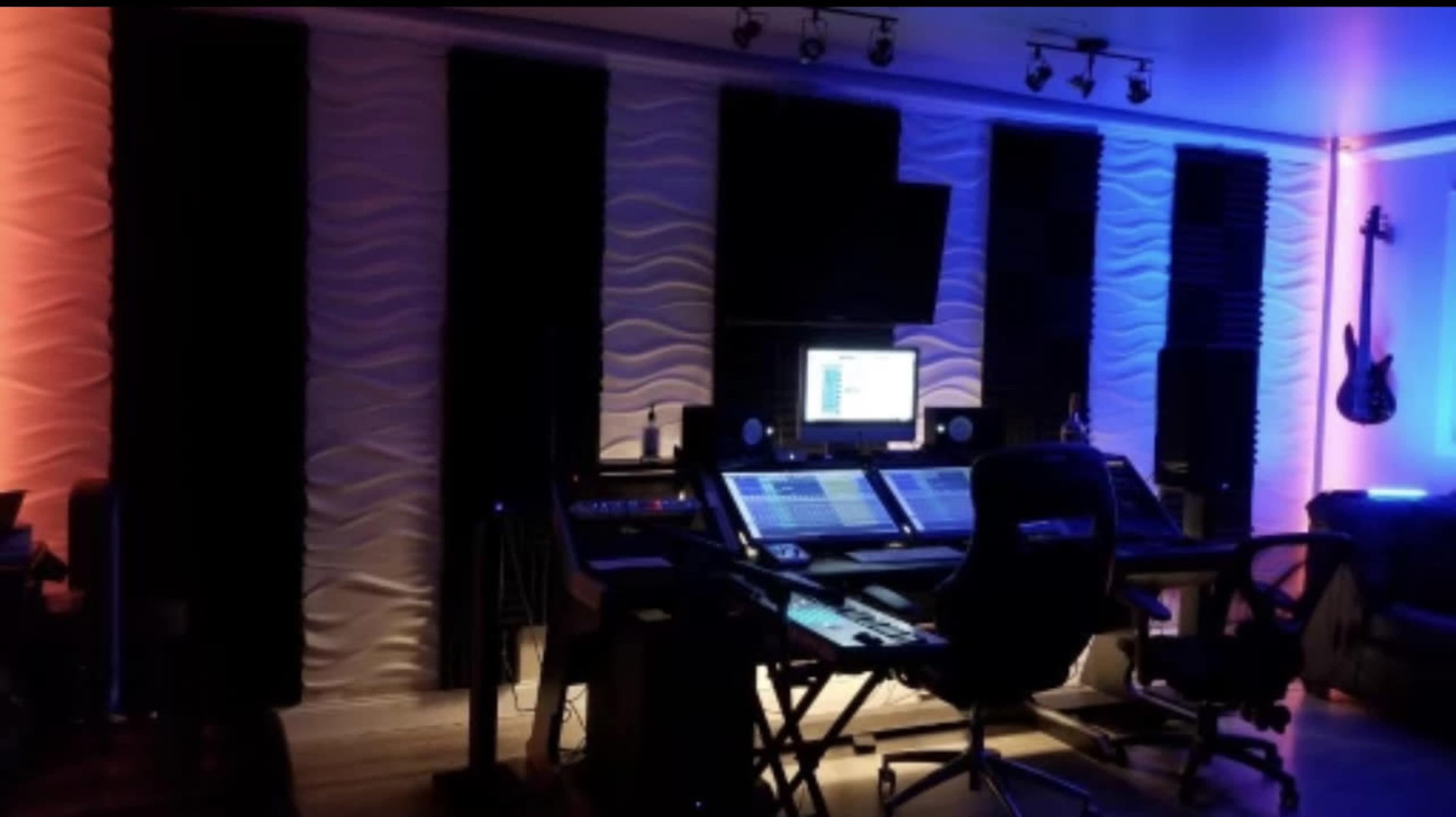 A music production studio with soundproof walls, a control desk equipped with audio equipment, and soft, colored lighting.