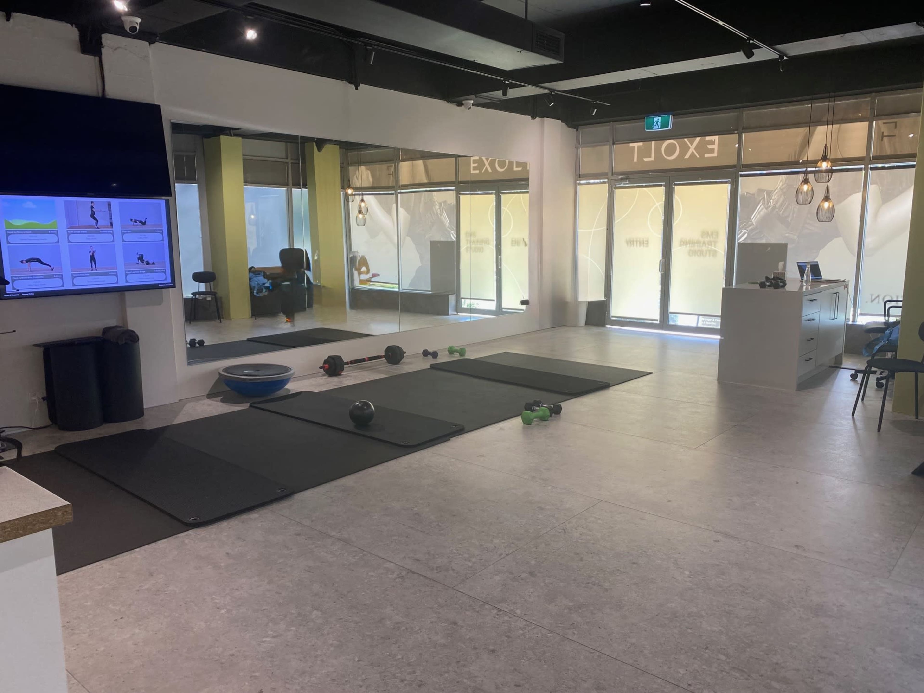 The image shows a fitness studio with exercise mats arranged on the floor, a large mirror, a television displaying workout instructions, and natural light coming through large windows.