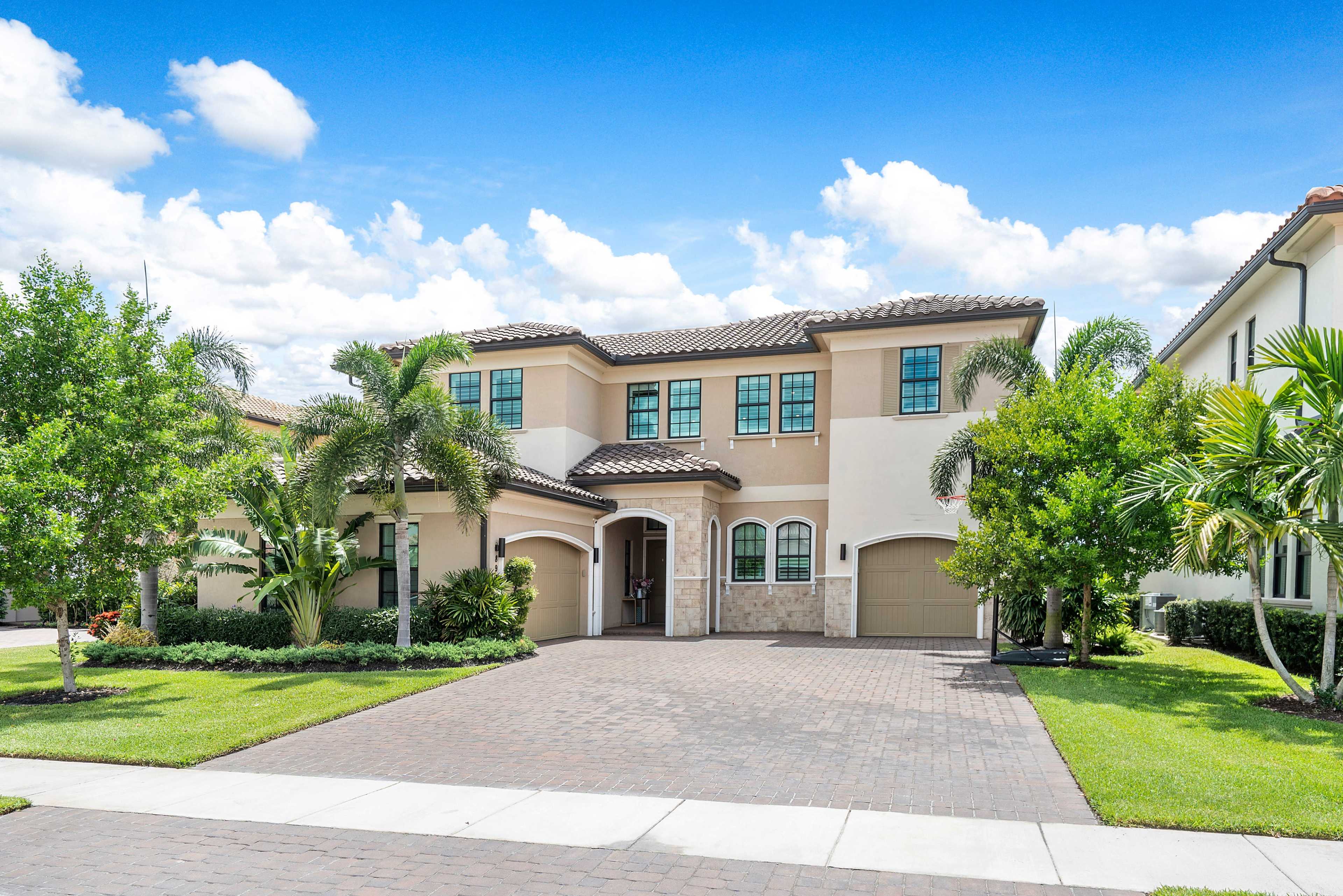 A two-story house with arched windows and a stone accent wall sits on a landscaped front lawn, featuring palm trees and a brick driveway.
