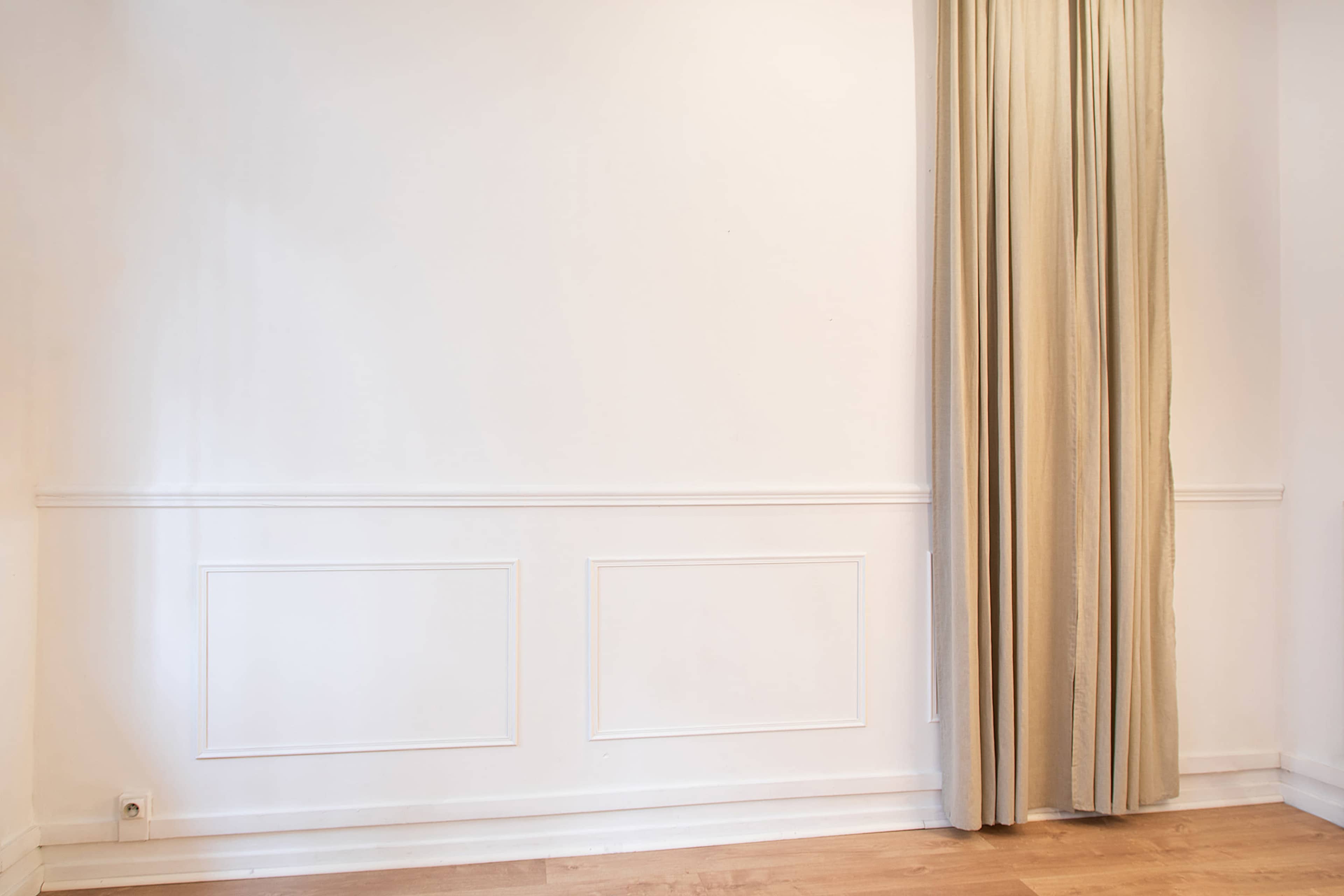A plain white wall with a decorative panel and a long, beige curtain on the right side.