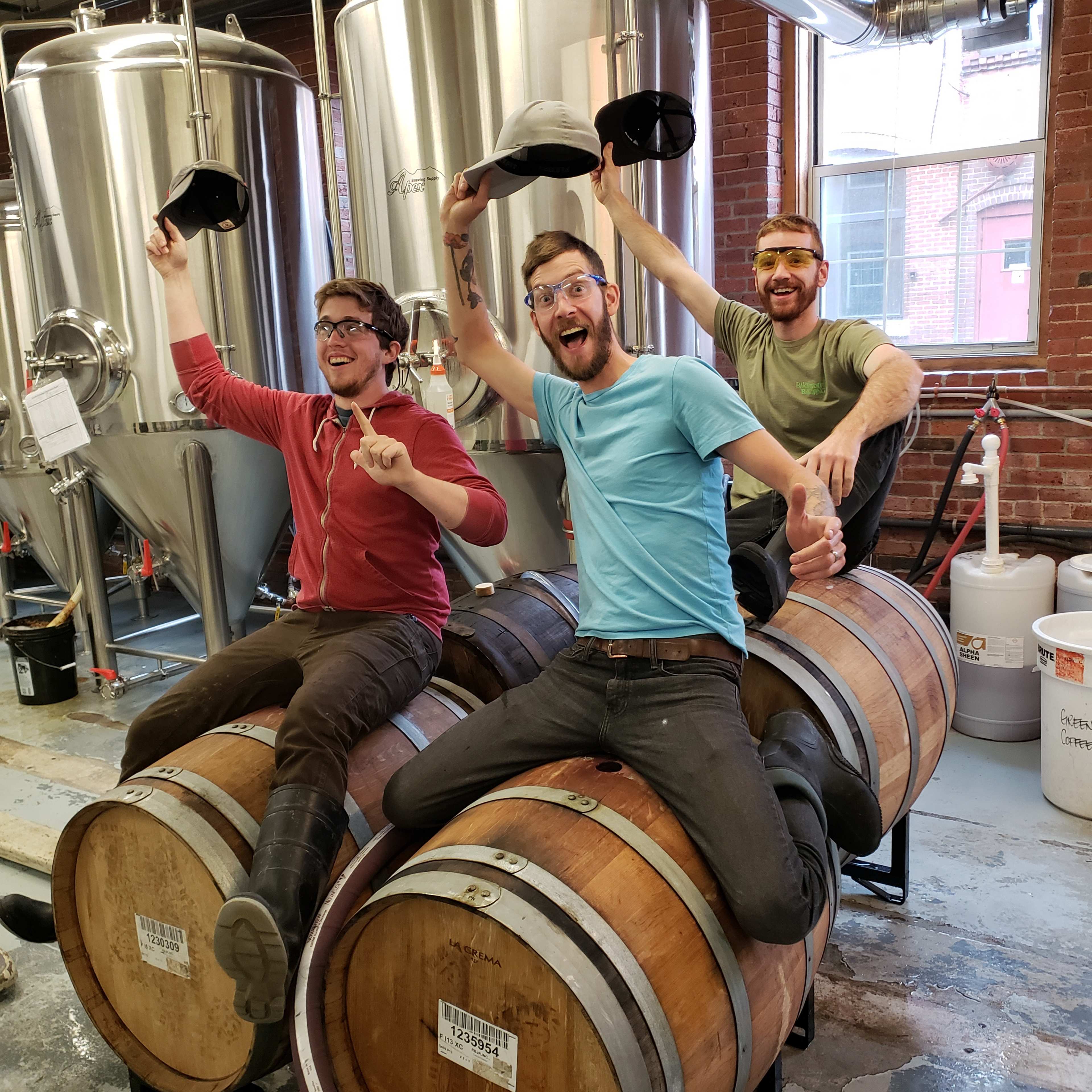 Three men sit on large wooden barrels in a brewery, celebrating and wearing hats while smiling and gesturing joyfully.