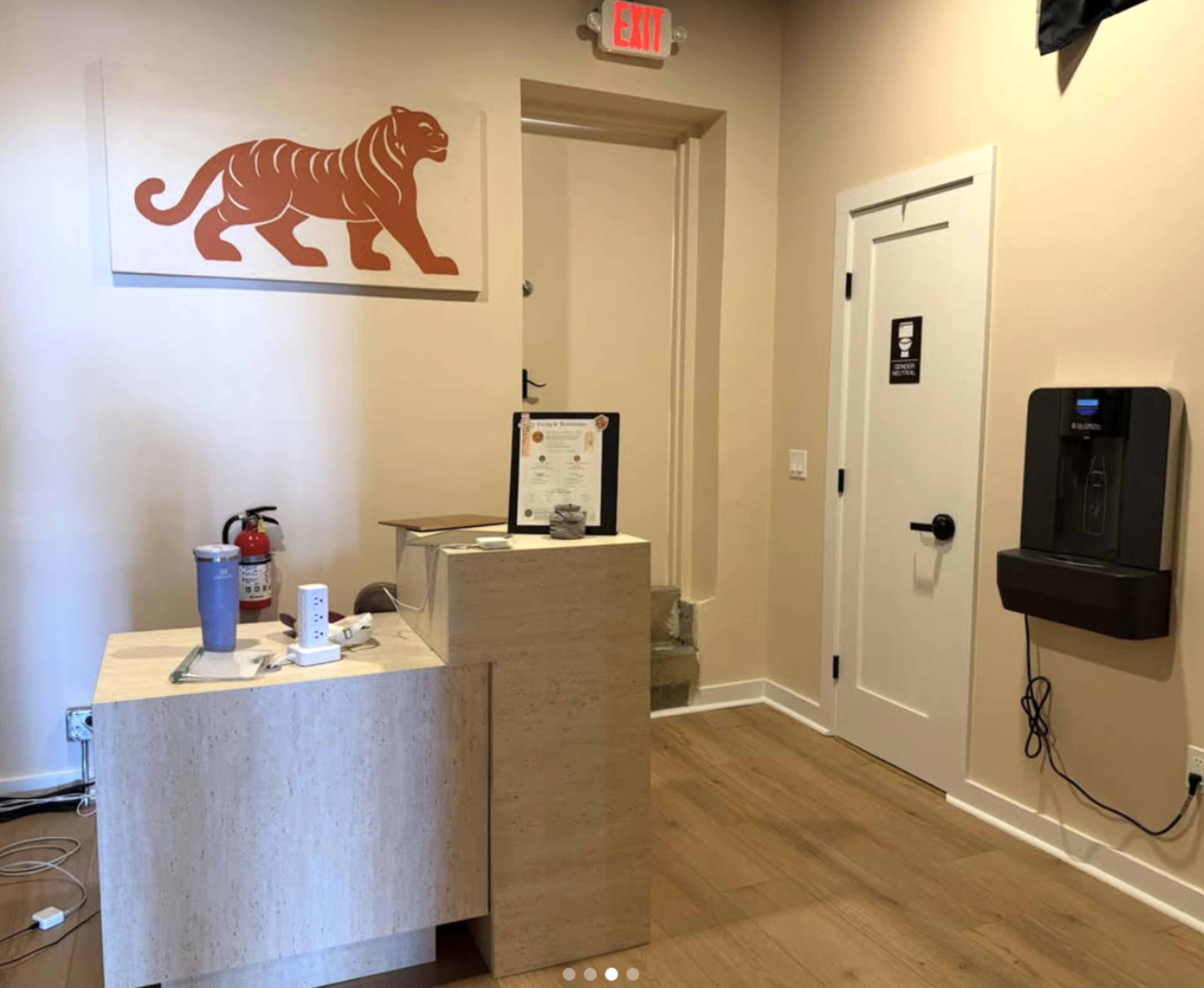 The image shows a small reception area with a wooden front desk, a fire extinguisher, a wall-mounted hand sanitizer dispenser, and a tiger artwork on the wall.