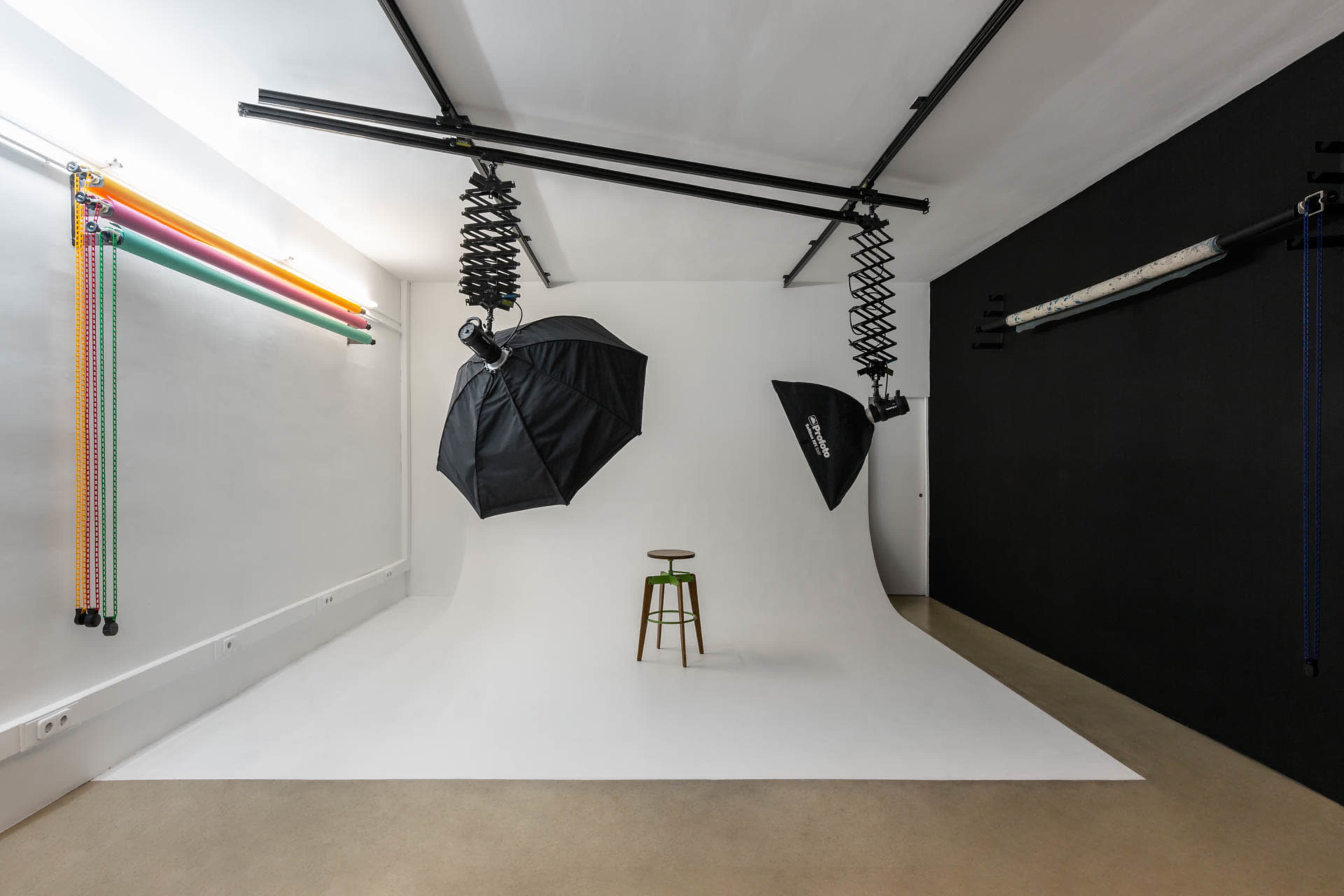 The image shows a photography studio with a white backdrop, a stool in the center, and various lighting equipment suspended from the ceiling.