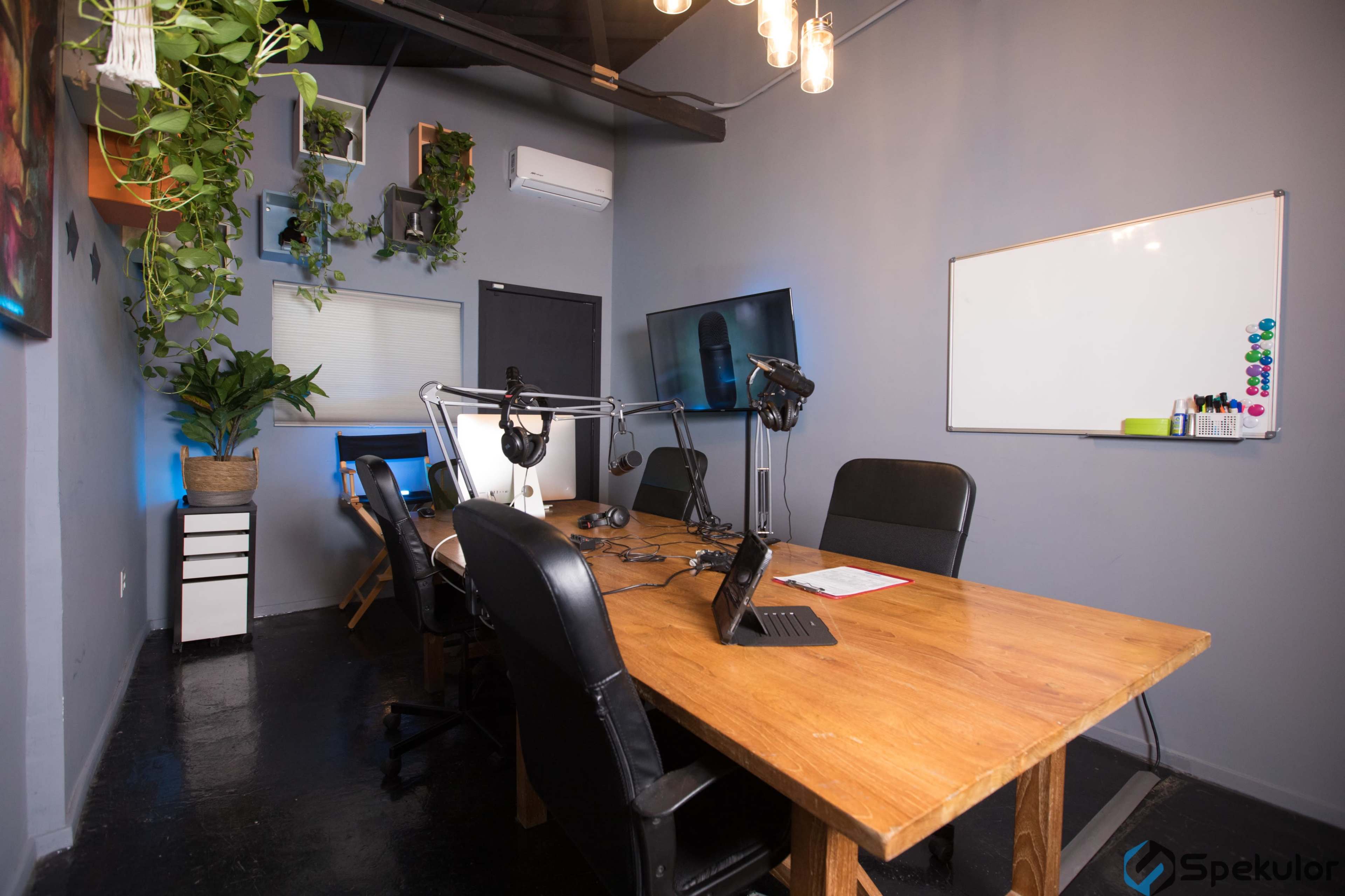 A modern conference room features a large wooden table surrounded by black chairs, equipped with microphones and computer screens, and adorned with plants and wall decorations.