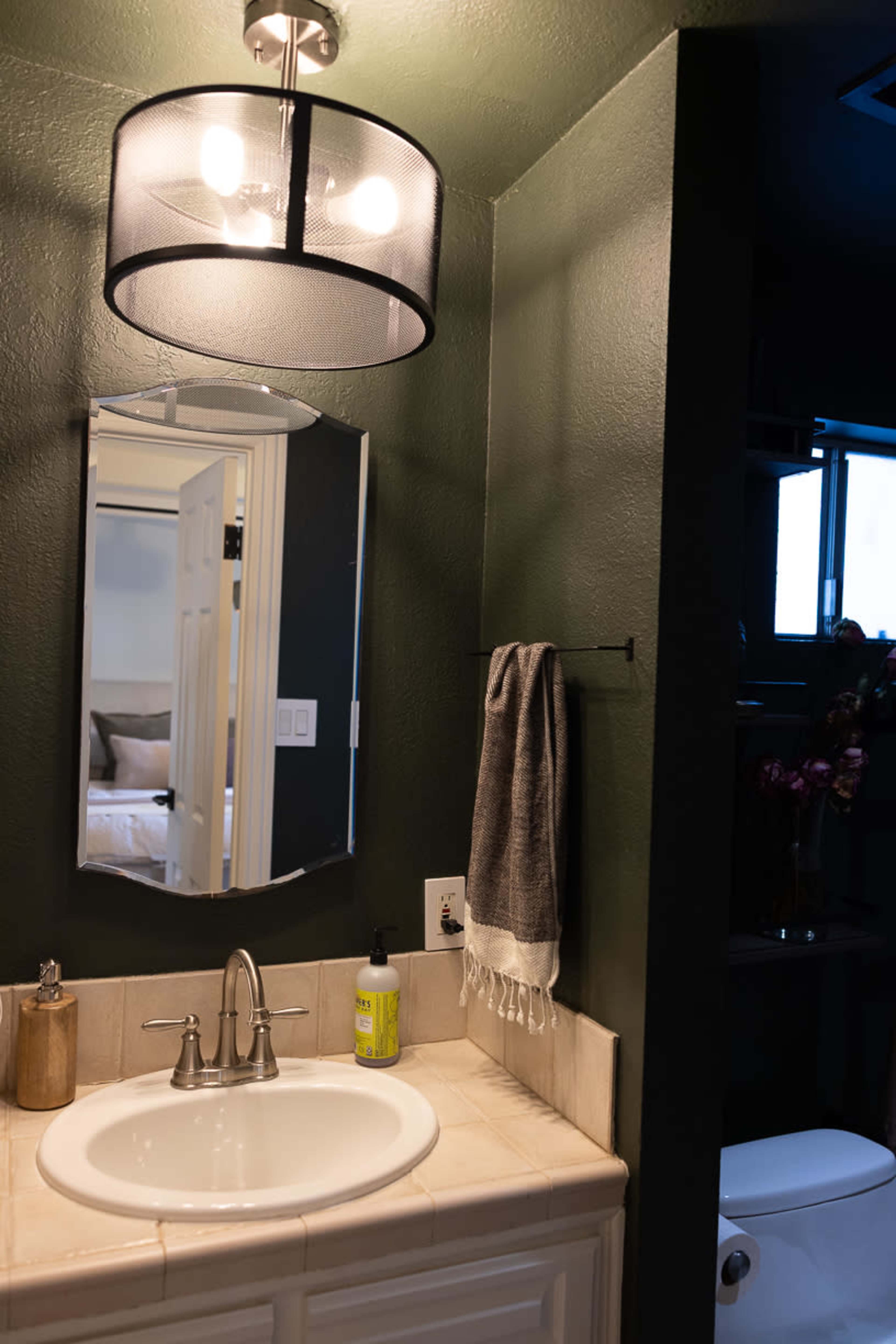 The image shows a bathroom with a circular light fixture, a large mirror above a sink, a hand towel hanging on a wall hook, and a toilet partially visible in the background.