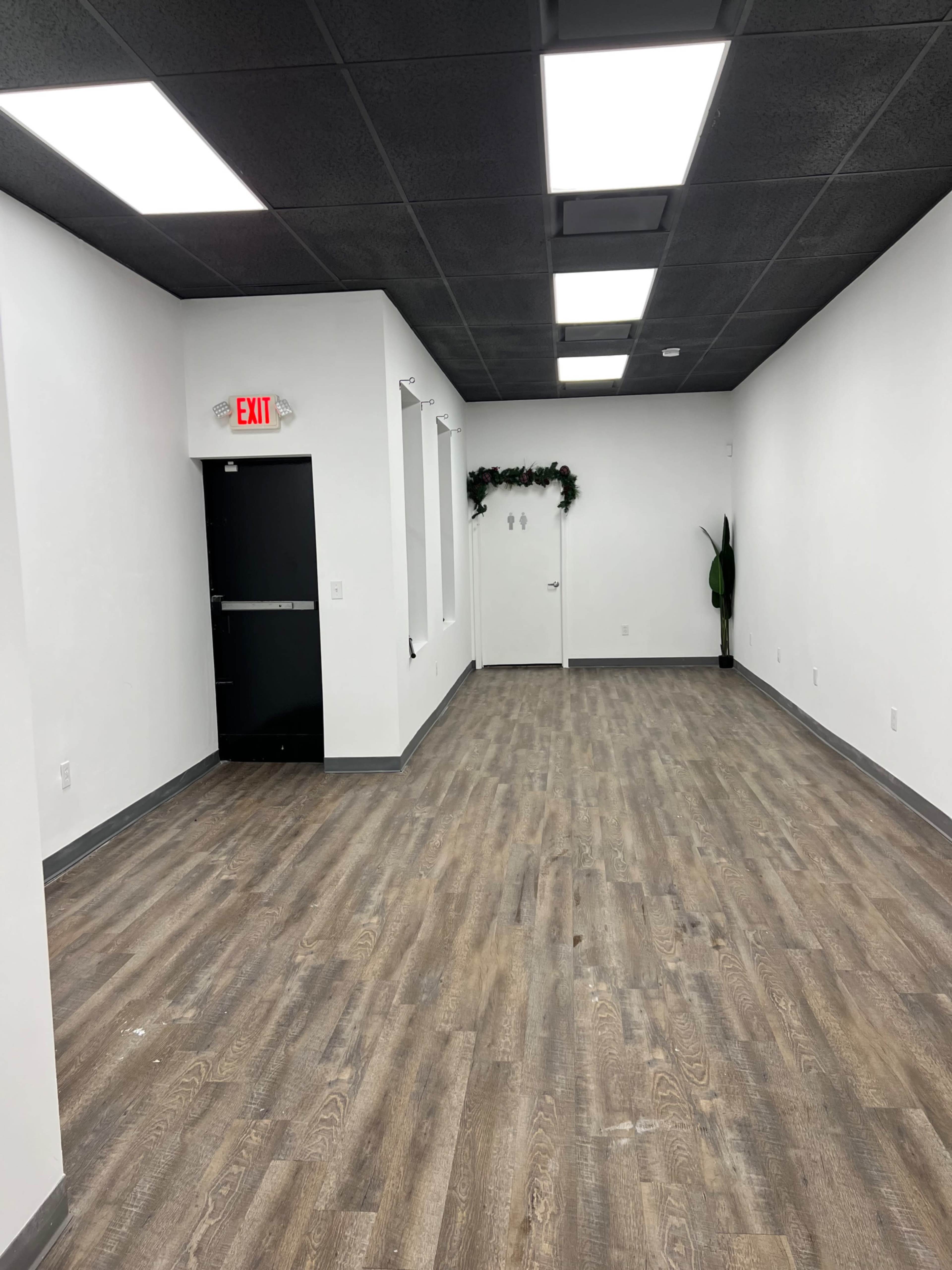 An empty room with wood flooring, white walls, and a door at the back, illuminated by bright overhead lights.