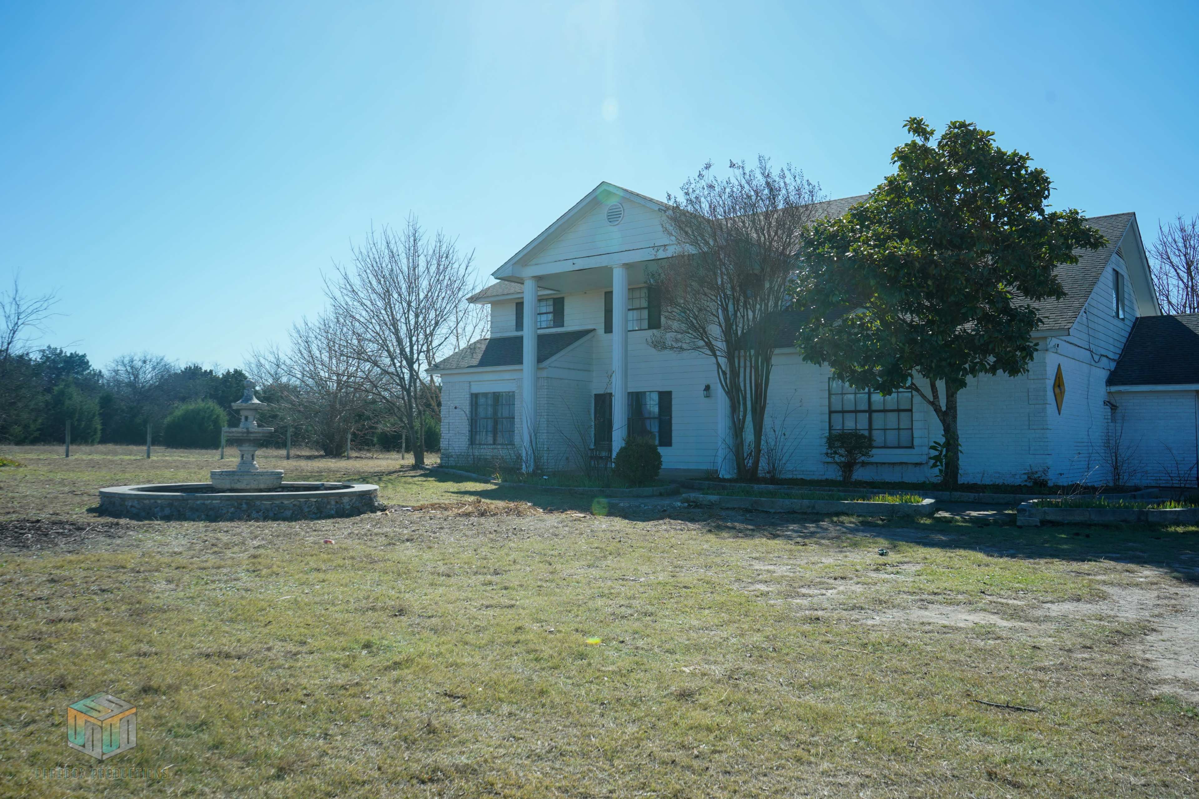 A large, two-story white house with a front porch is situated next to a fountain in an open grassy area.