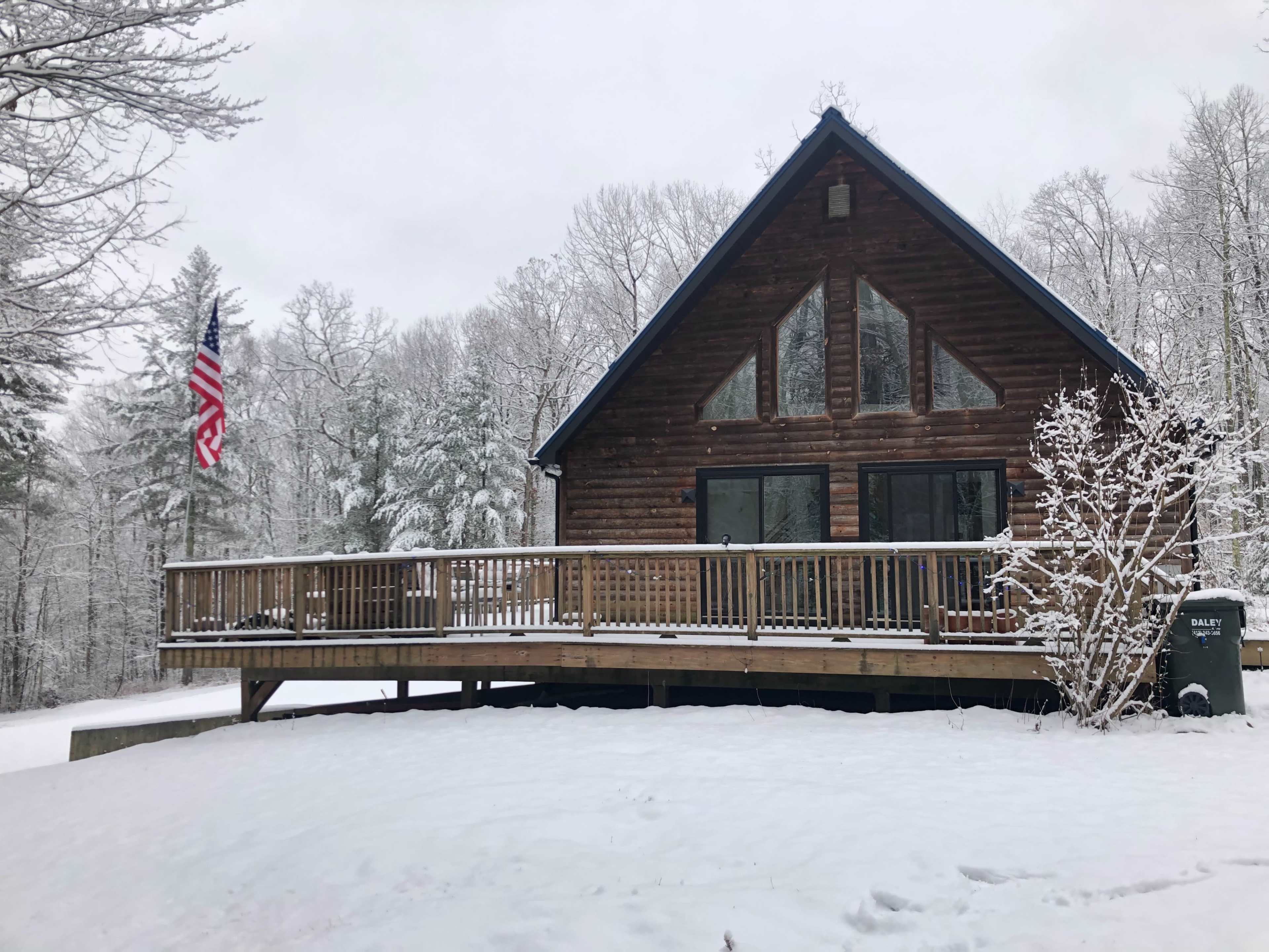 A wooden cabin with a deck is surrounded by snow-covered trees and a flagpole displaying the American flag.