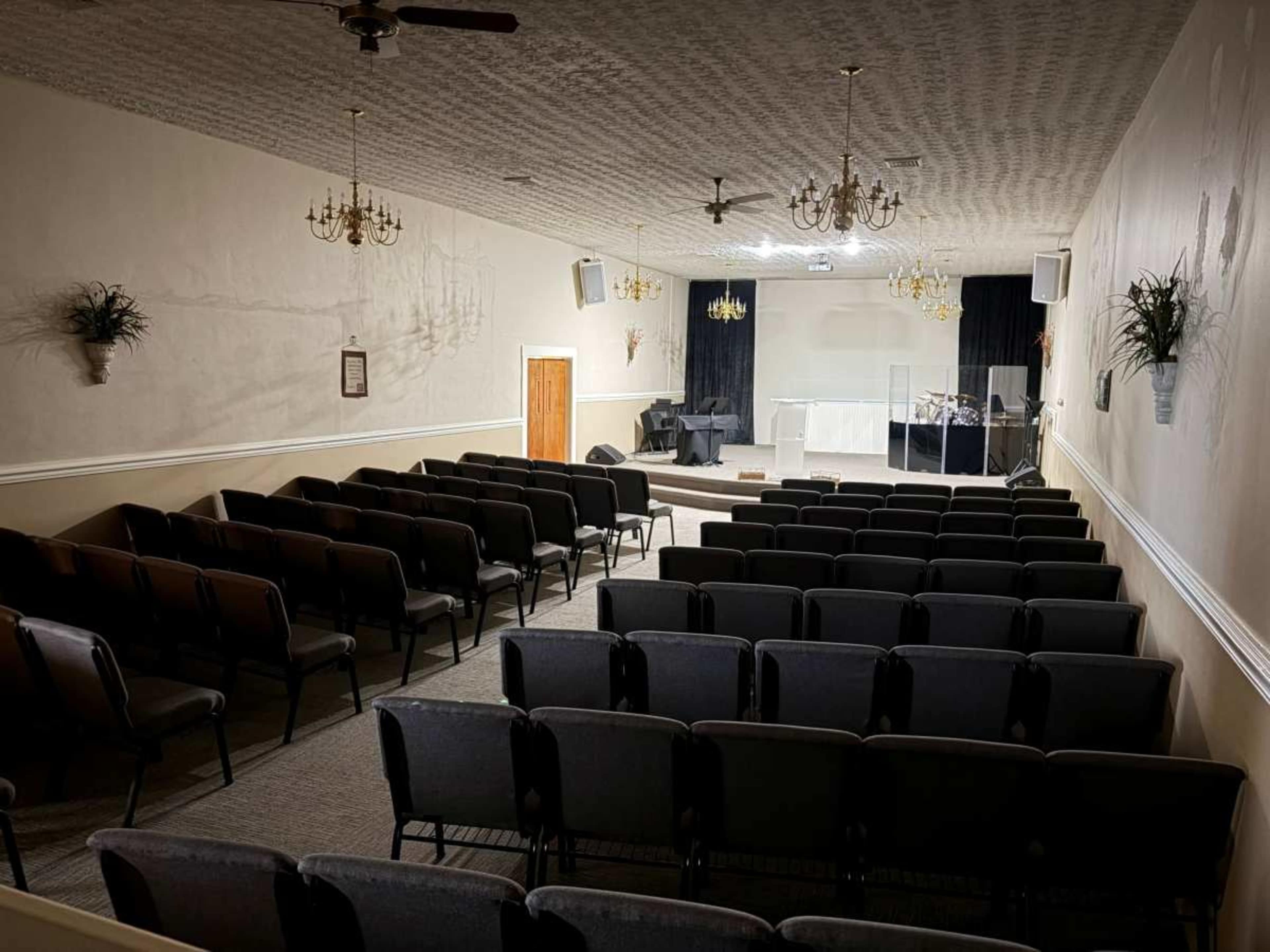 The image shows the interior of a church with rows of empty chairs facing a stage area, featuring decorative chandeliers and a simple backdrop.