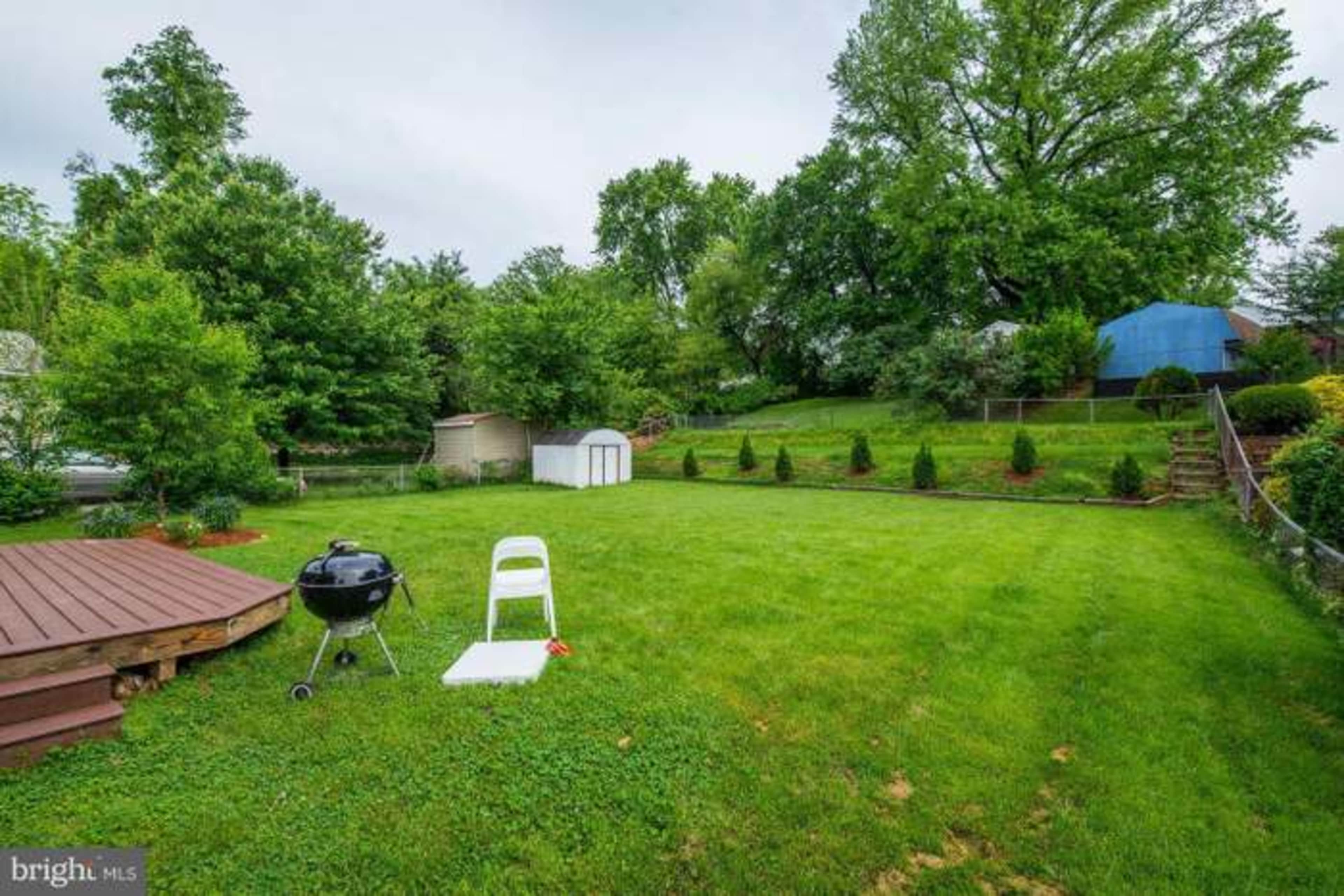 The image shows a grassy backyard with a charcoal grill, a white folding chair, a small shed, and trees in the background.