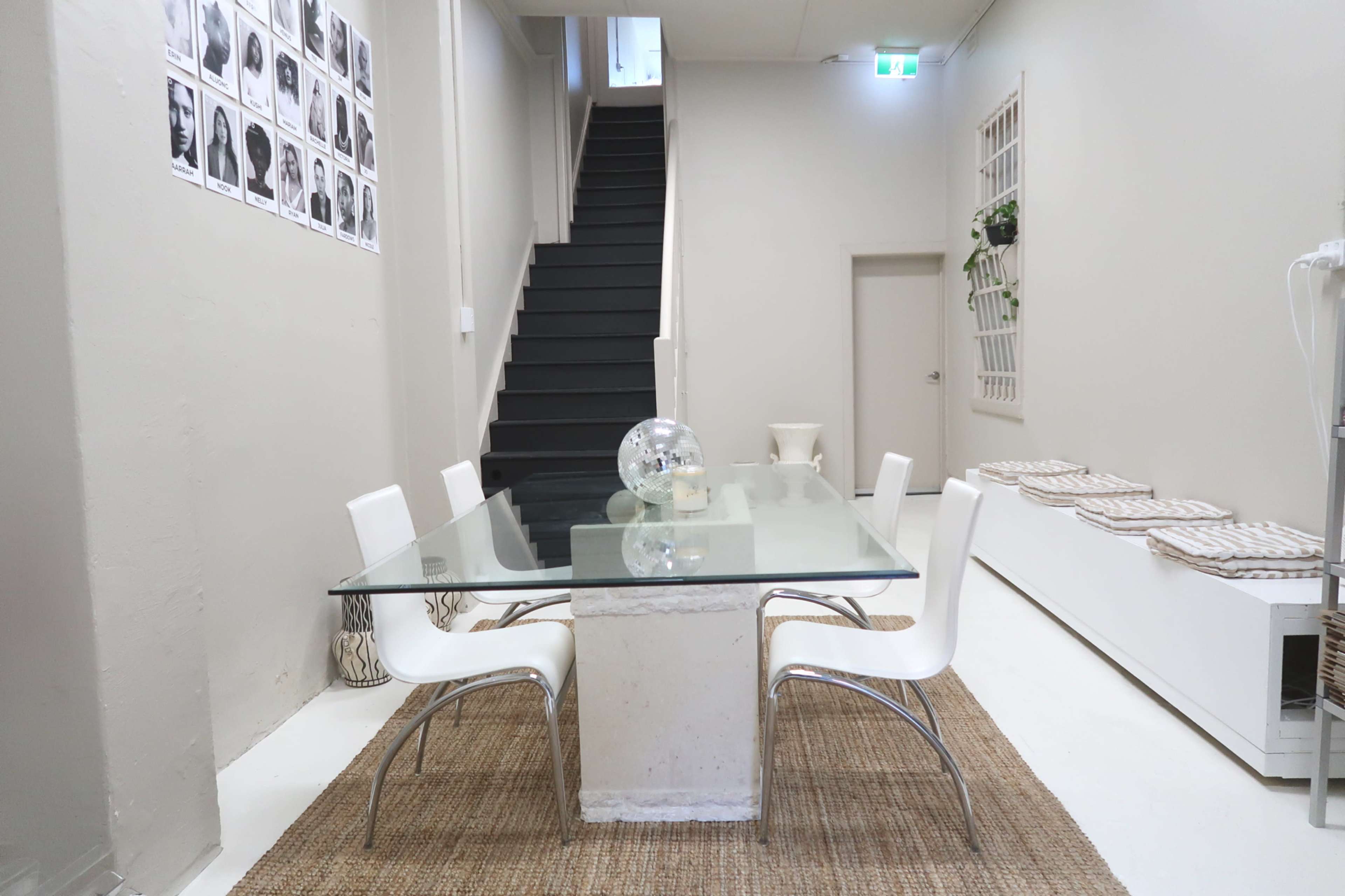 A glass dining table with white chairs is positioned on a woven rug, alongside a staircase and a wall adorned with black and white photographs.