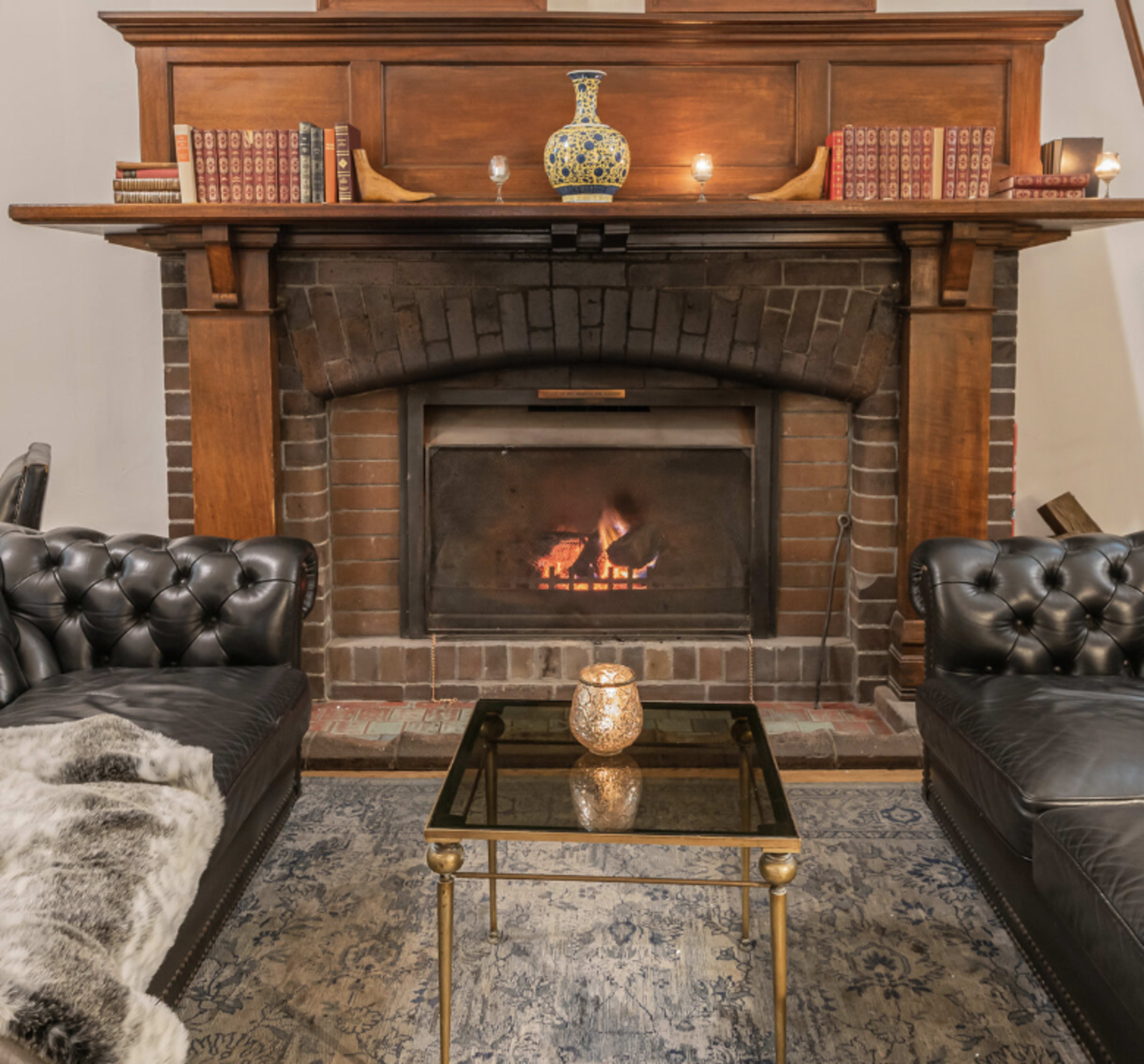 A cozy living room features a brick fireplace with a fire burning, flanked by two black leather couches and a glass coffee table.