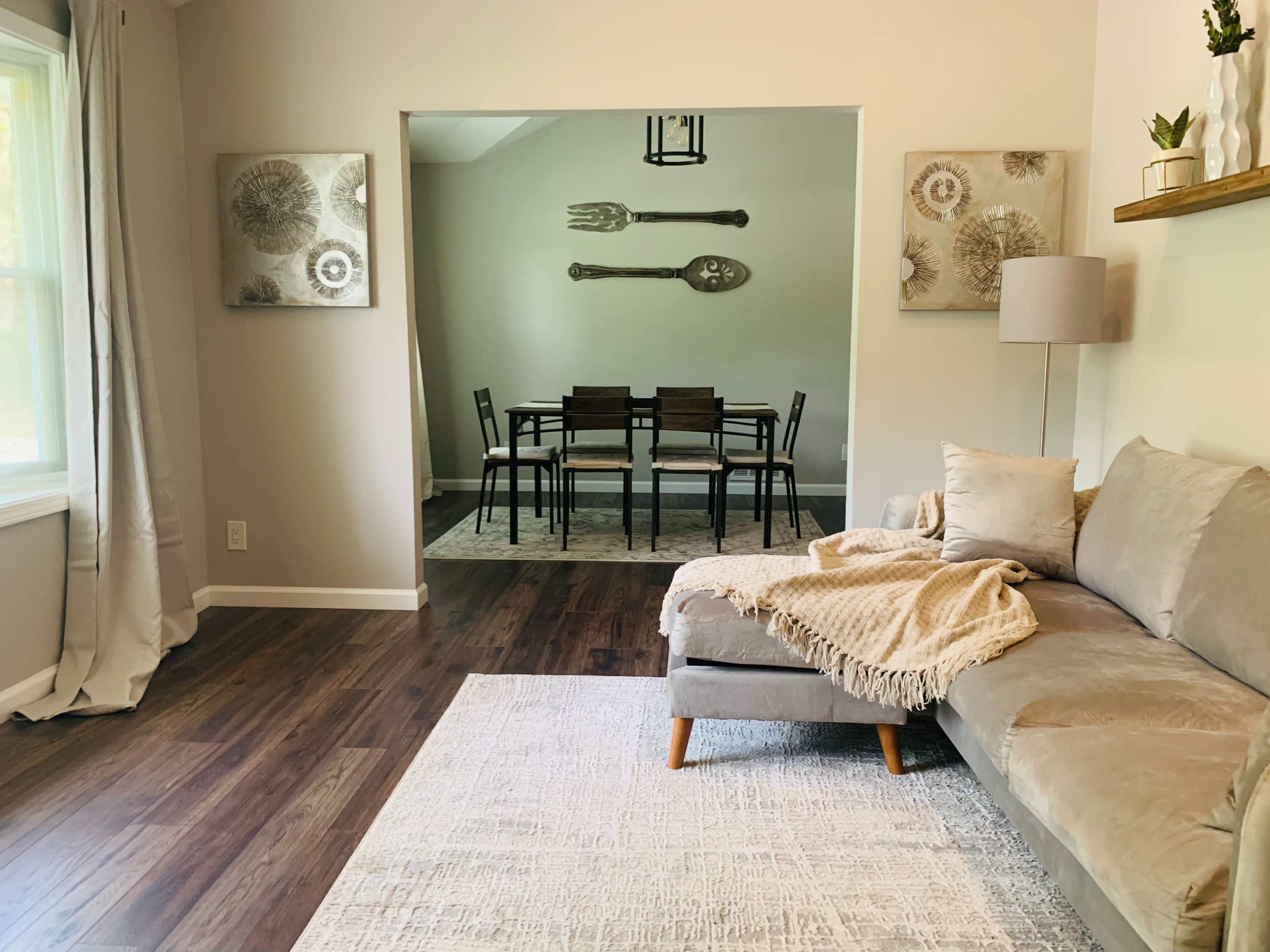 A light-filled living room features a gray sectional sofa with a blanket, leading into a dining area with a table and chairs, adorned by wall art depicting kitchen utensils.