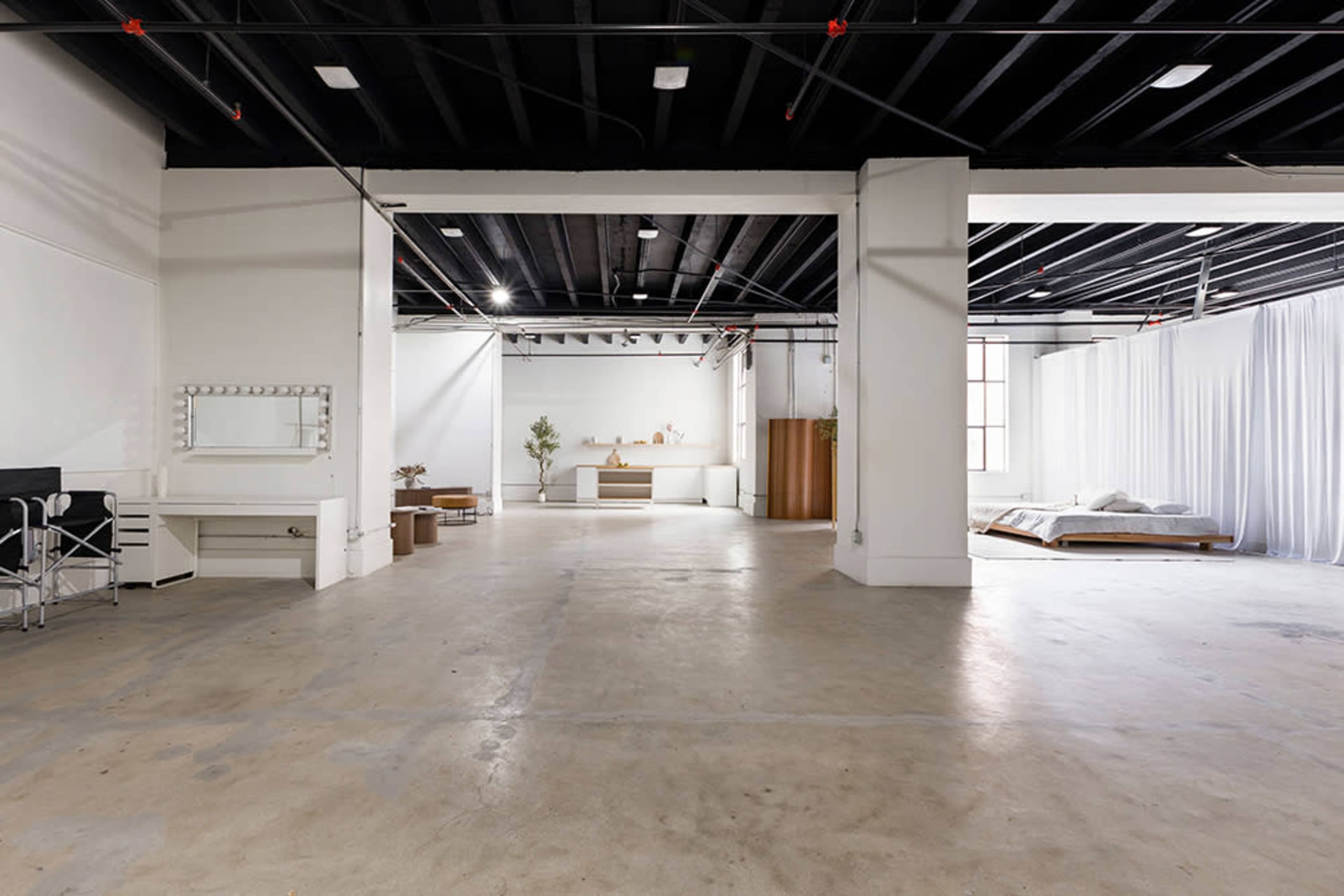 The image shows a spacious, empty interior with polished concrete flooring, white walls, and a minimalist design featuring a bed, a dressing table with lights, and draped fabric partitions.