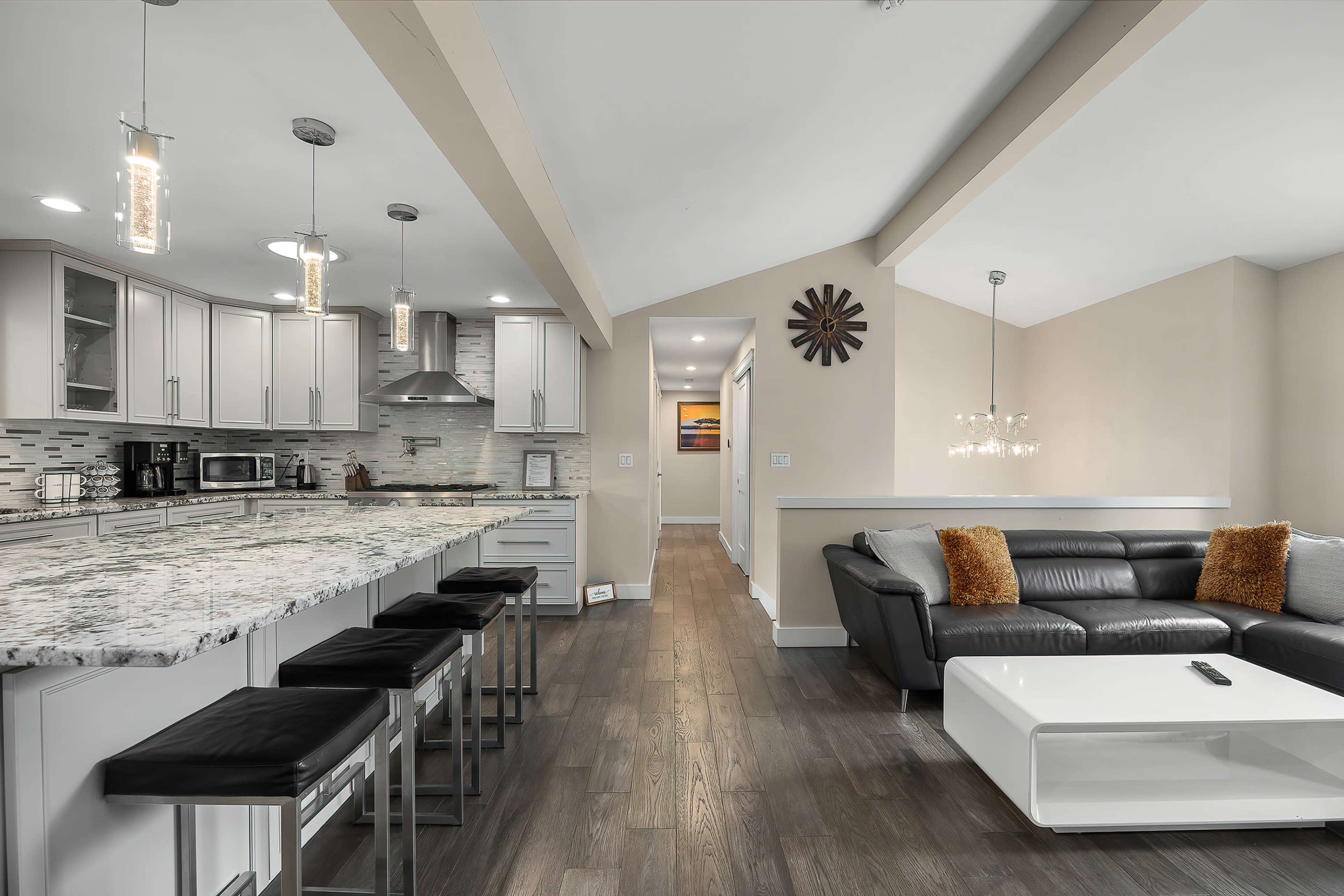 A modern kitchen and living area with a large marble island, stainless steel appliances, and a black leather couch facing a minimalist coffee table.
