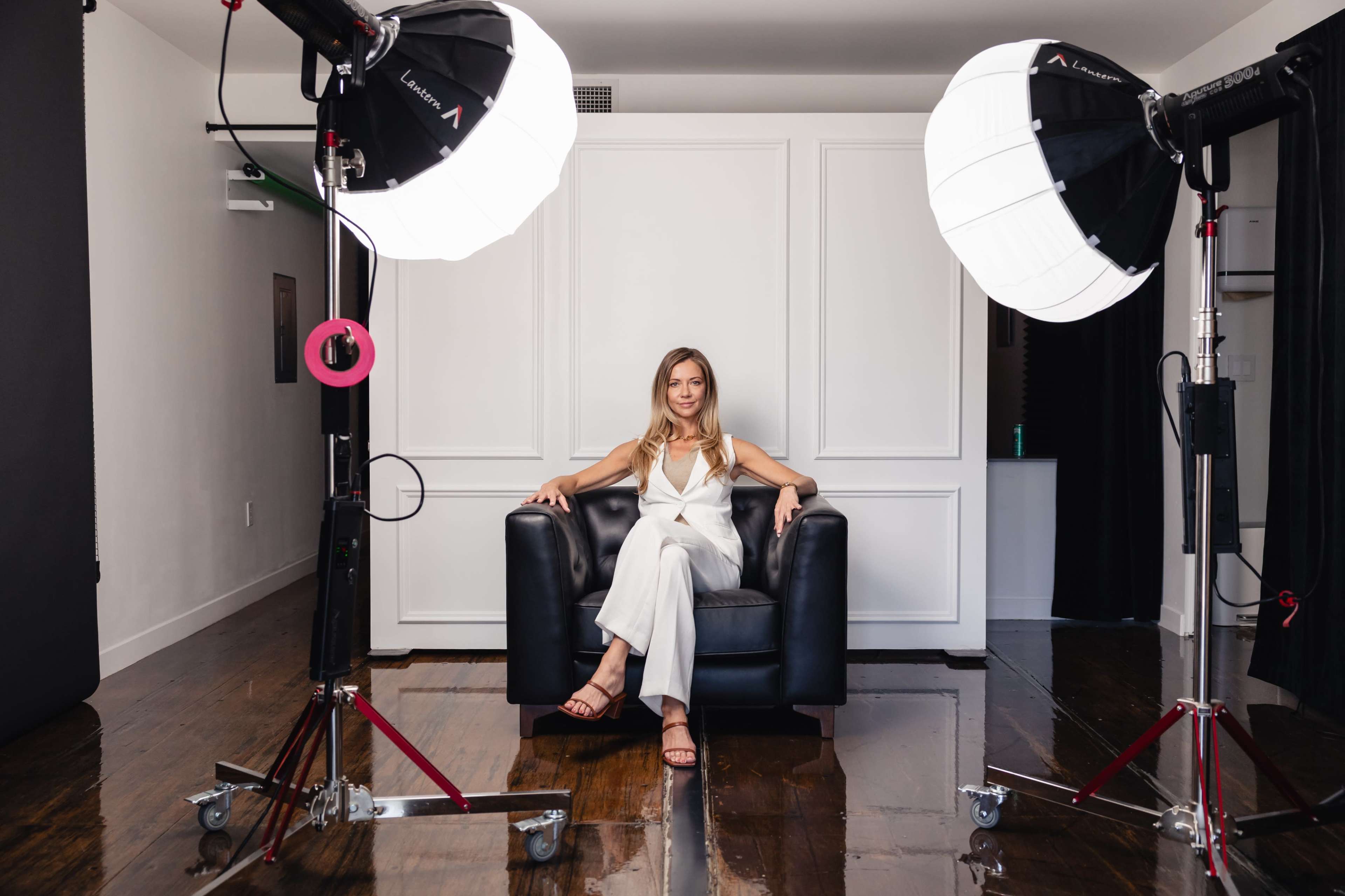 A woman sits confidently in a black chair between two large softbox lights in a photography studio.
