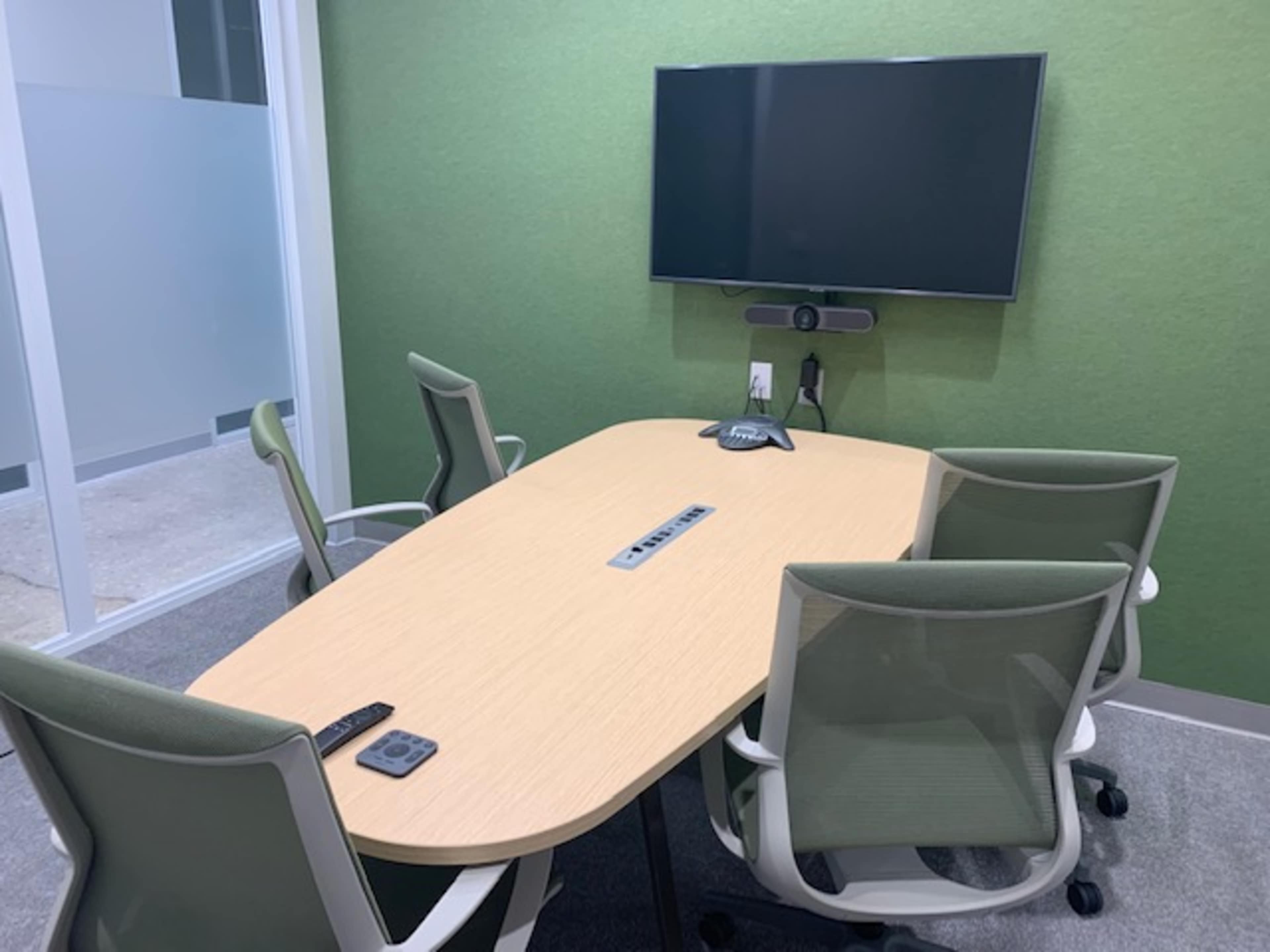 A small, modern conference room with a green wall, an oval table, and four ergonomic chairs, along with a mounted television at one end.