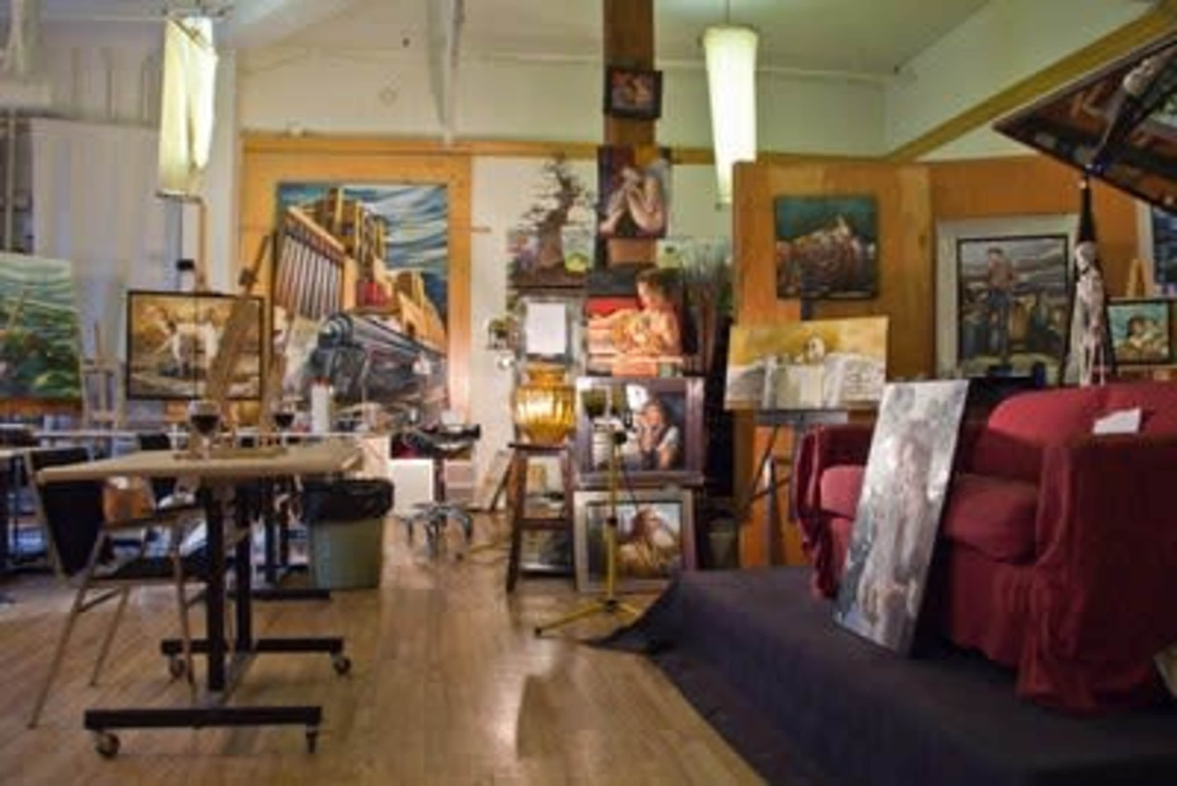 An artist's studio filled with various paintings, an easel, and a red sofa, with natural light illuminating the space.