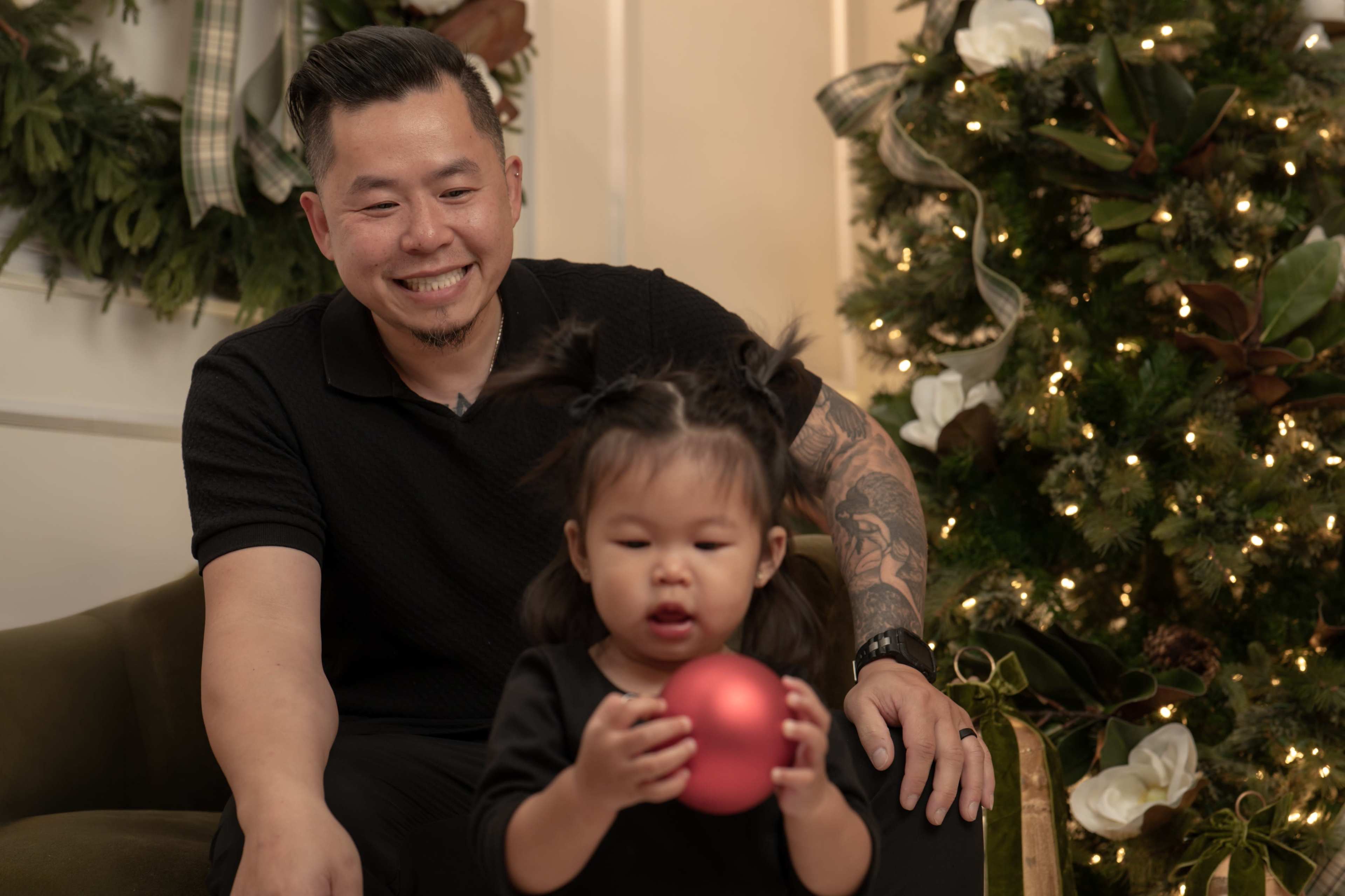 A man sits on a couch beside a young girl who is holding a red ornament, in front of a decorated Christmas tree.