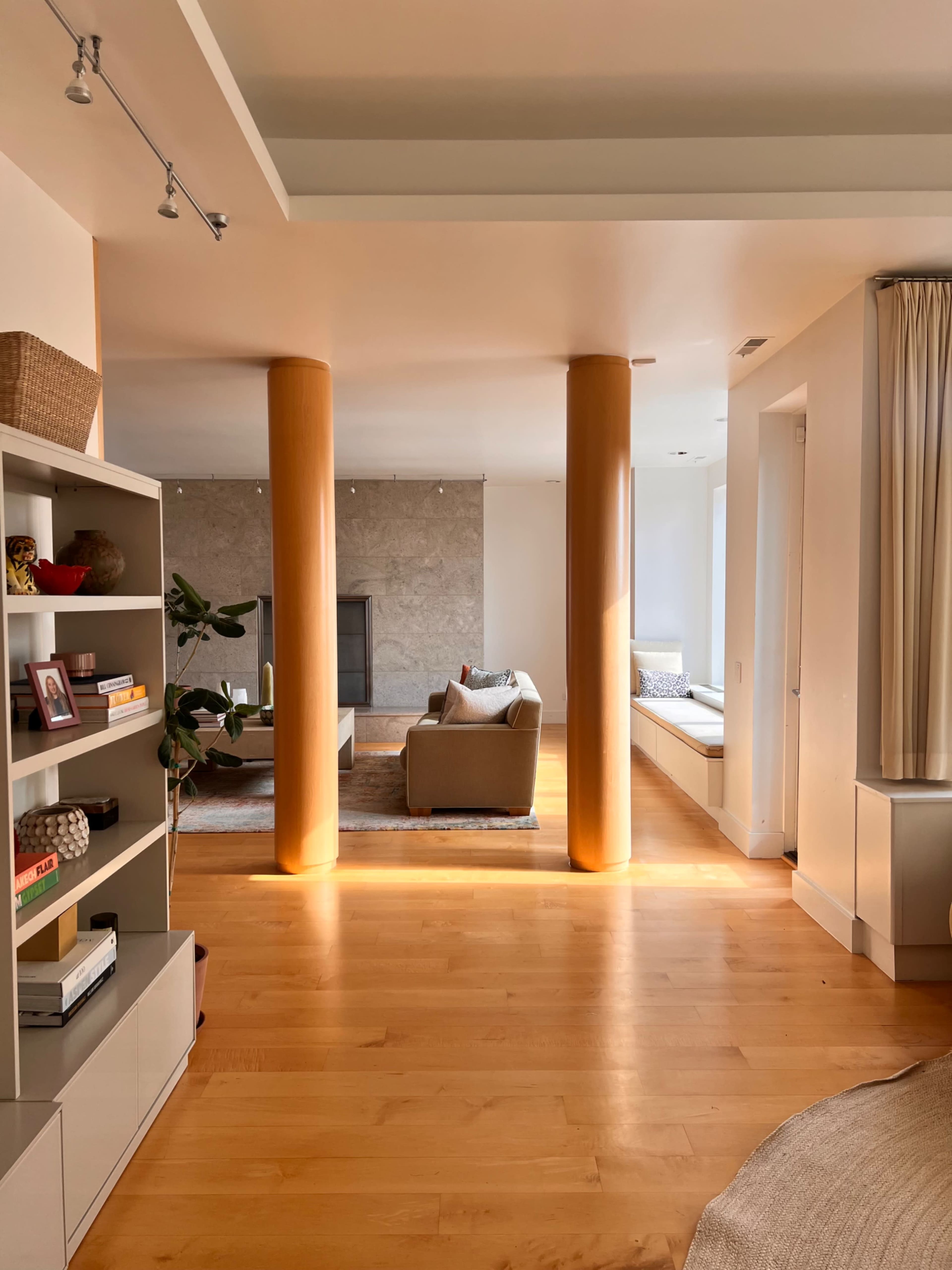 The image shows a well-lit, modern living space with wooden flooring, featuring a shelf, two vertical columns, and a cozy seating area.