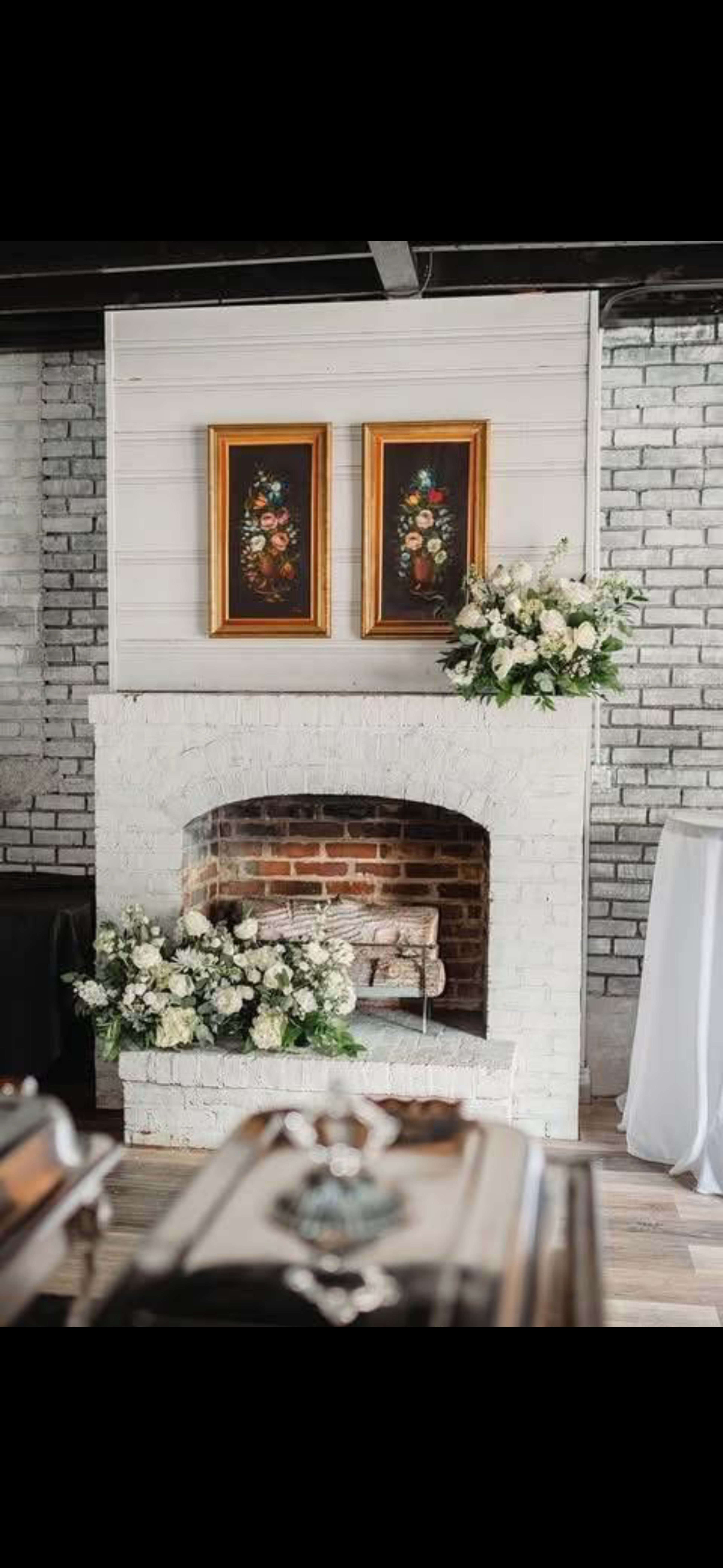 The image shows a white-brick fireplace with floral arrangements on the mantel and beside it, framed paintings hanging above.