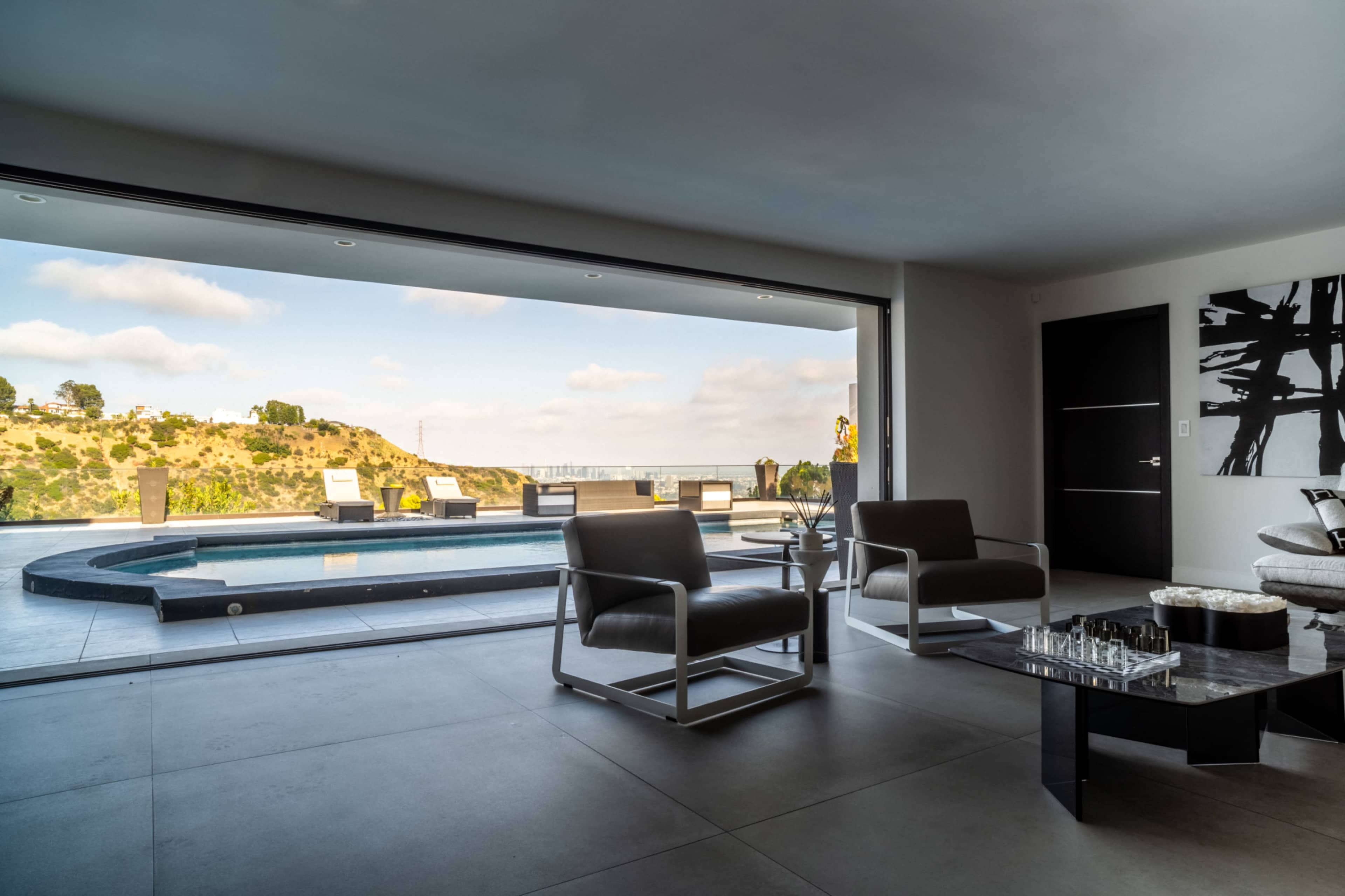 A modern living room opens to a pool area with a scenic view of hills and lounge chairs.