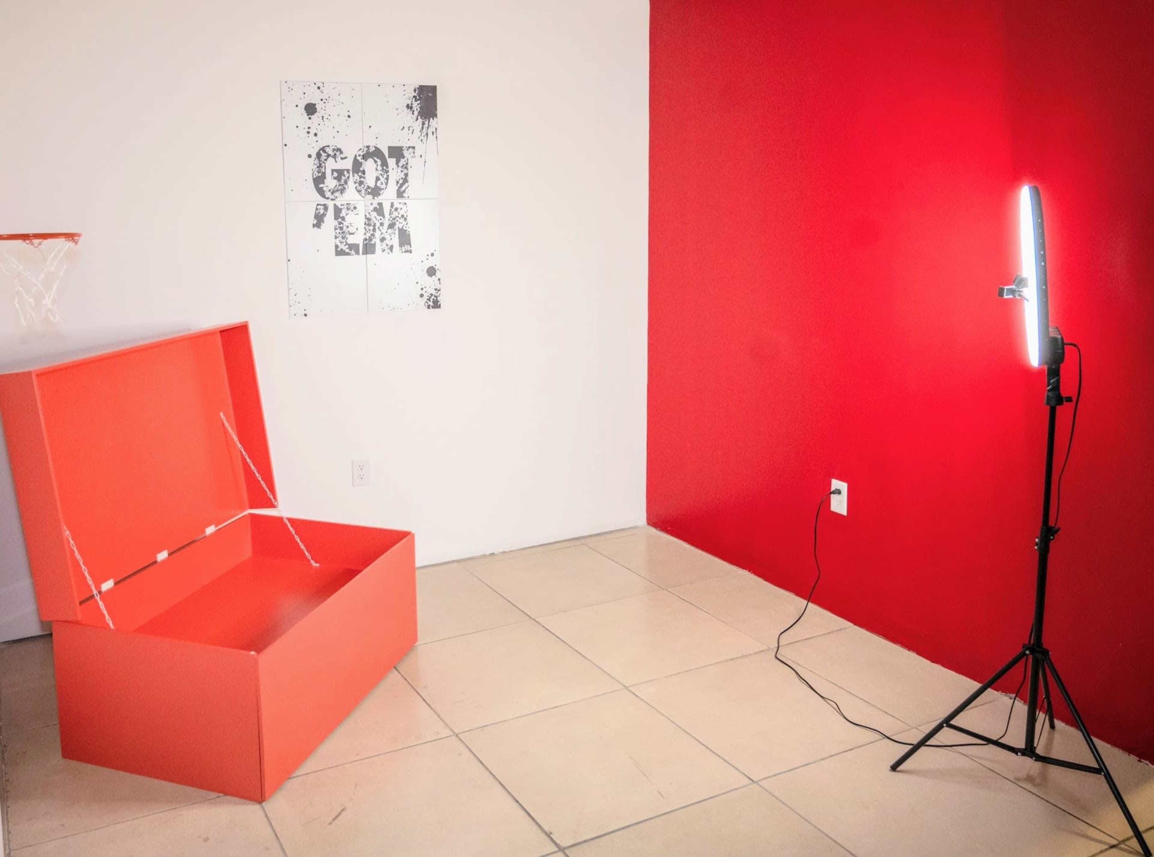A room with a red wall, a large orange box in the center, a basketball hoop on the left, and a standing light fixture on the right.