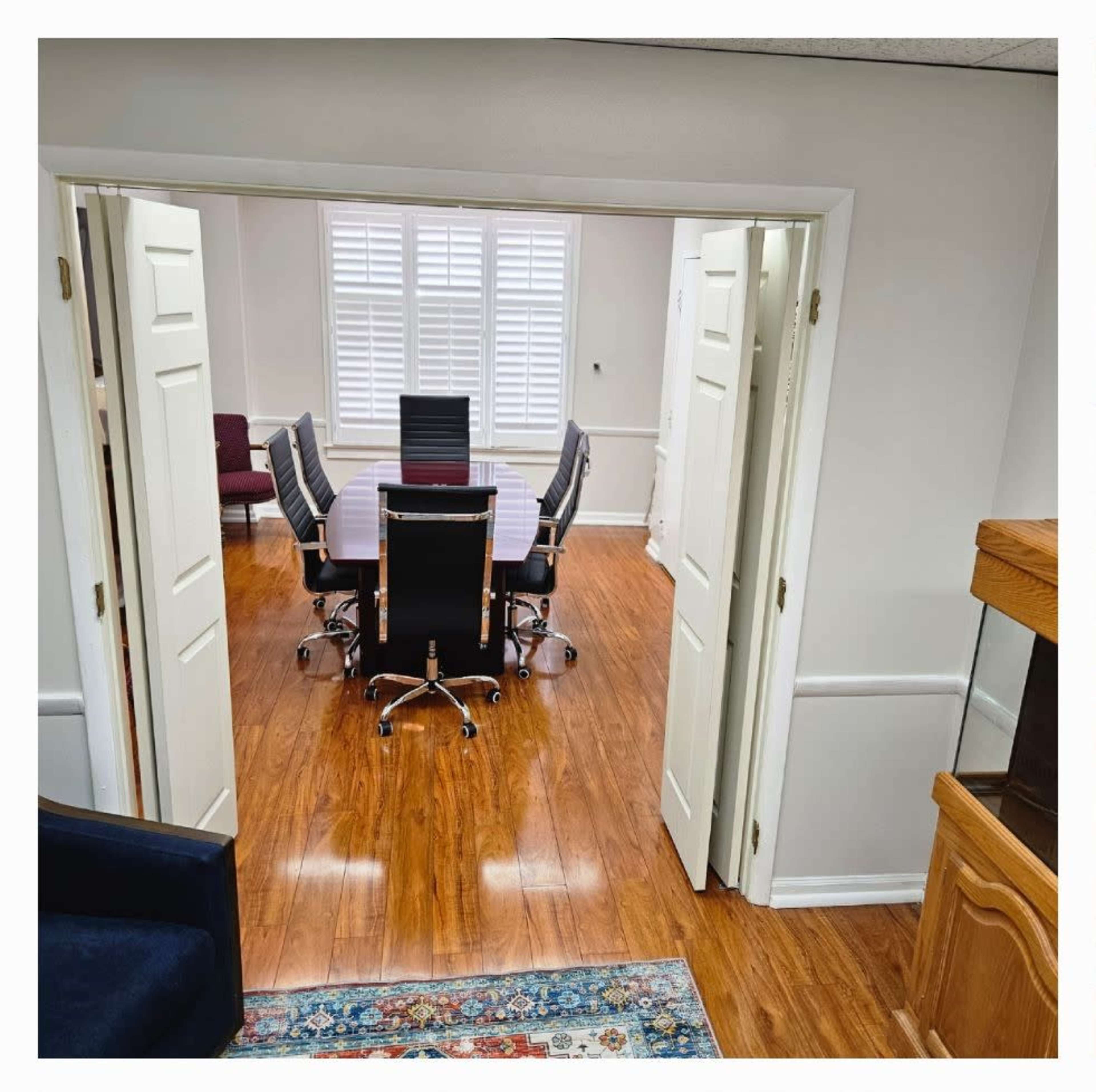 The image shows a view of a conference room through an open doorway, featuring a large table surrounded by black chairs and bright lighting.