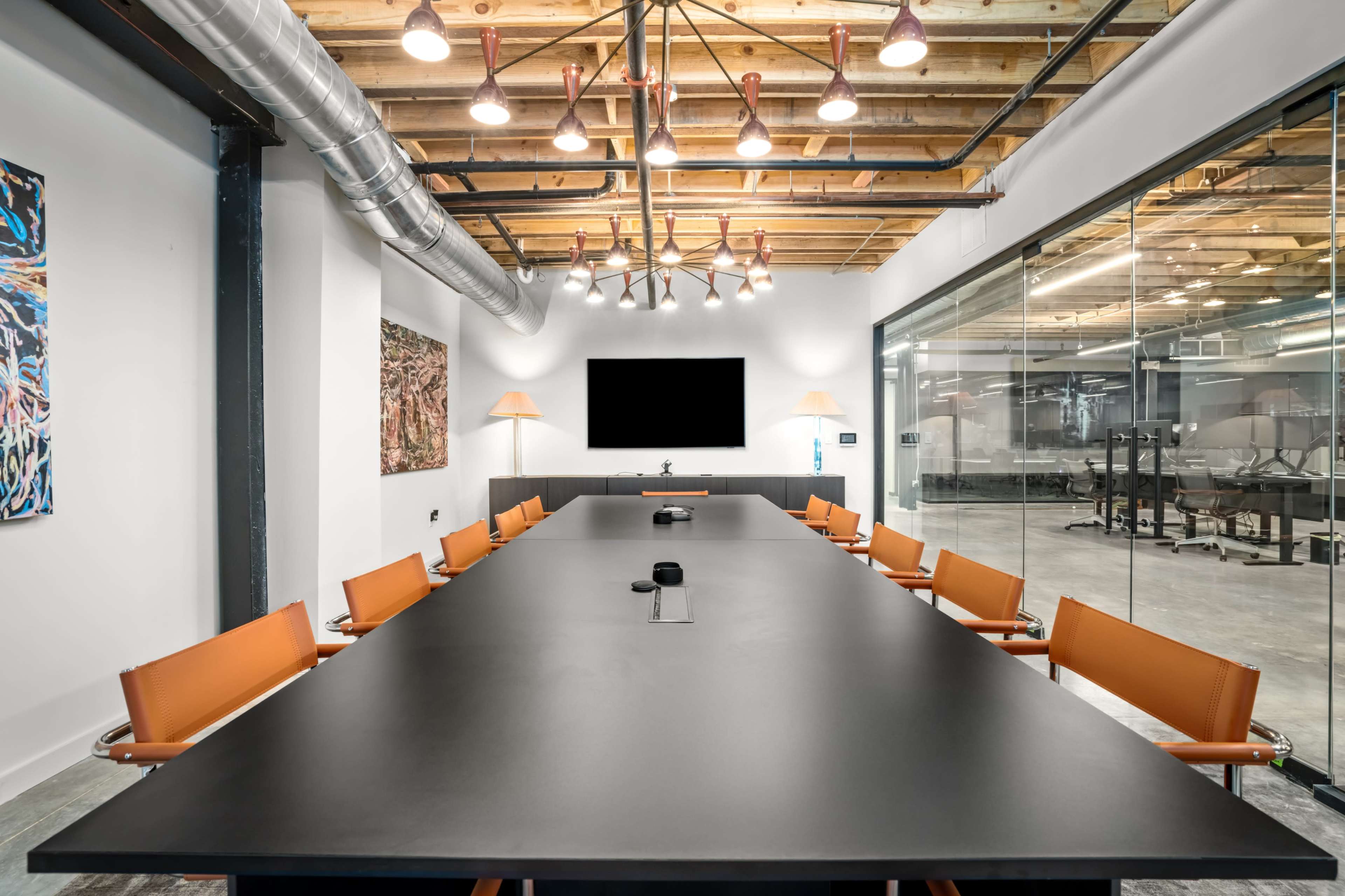 A modern conference room features a long, dark table with orange chairs, a wall-mounted screen, and large glass walls showcasing the surrounding space.
