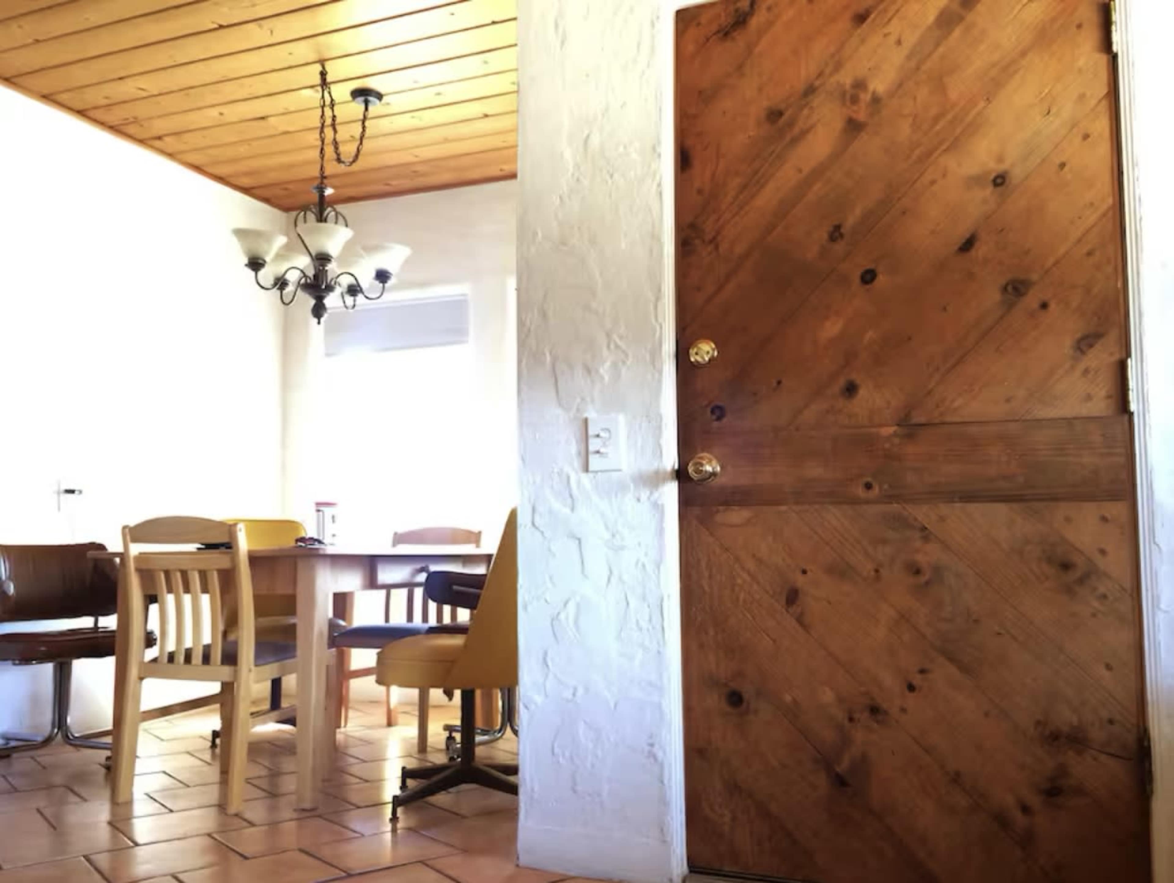 The image shows a wooden door with diagonal paneling leading into a dining area furnished with a table and chairs under a chandelier.