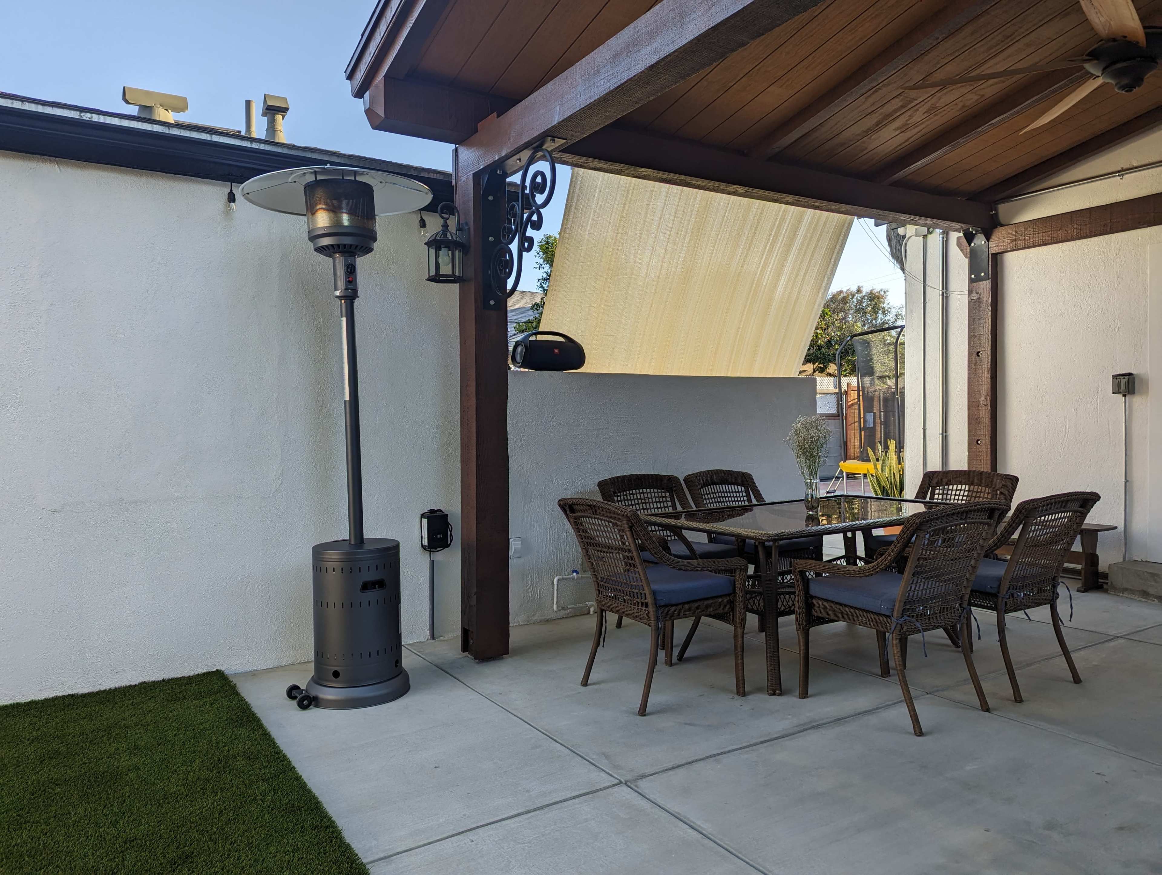 A patio area with a dining table and chairs under a wooden pergola, accompanied by a patio heater and a wall-mounted light fixture.