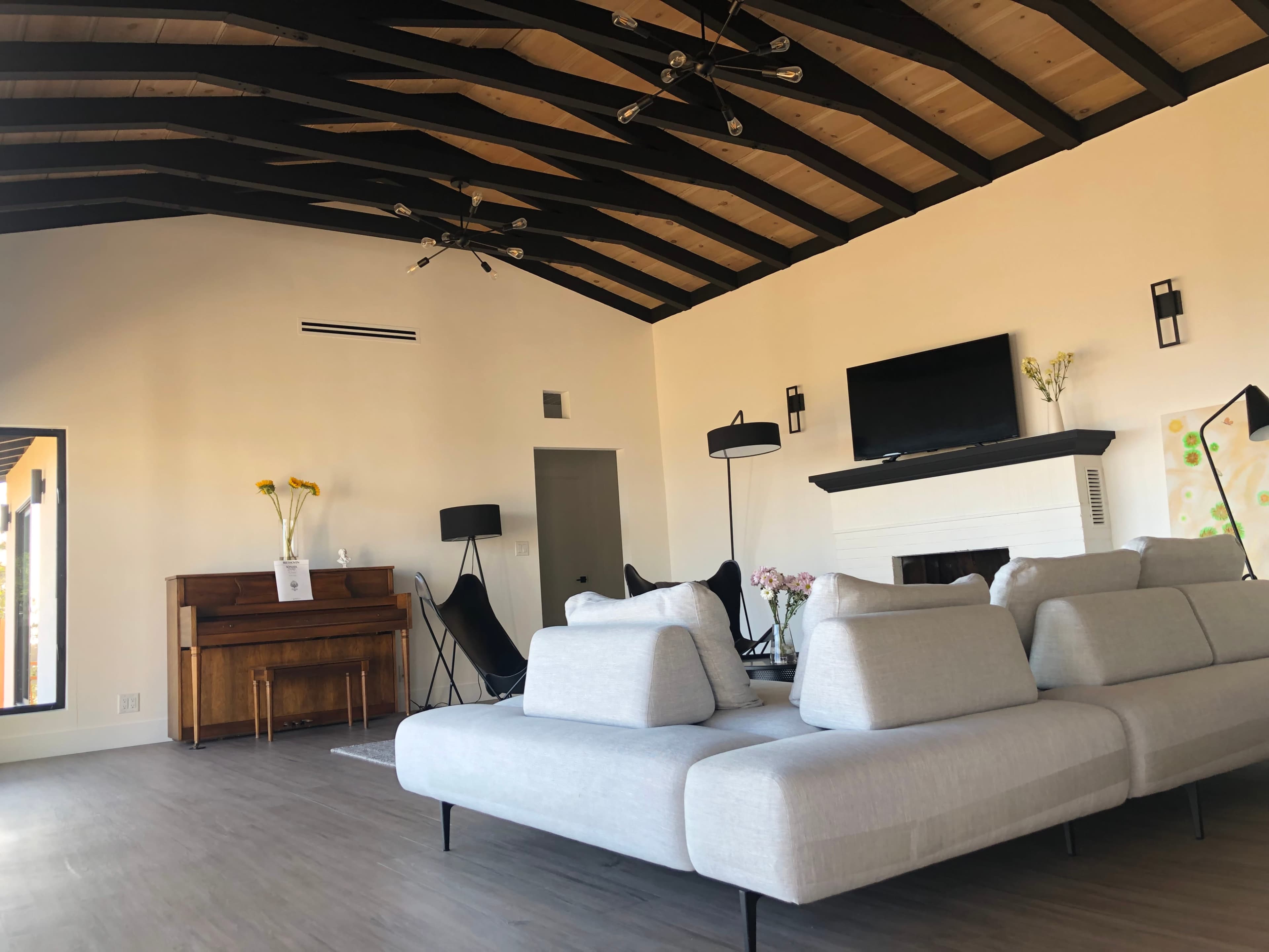 A modern living room with a large sectional sofa, a piano, and a television mounted above a fireplace, featuring high ceilings with exposed wooden beams.