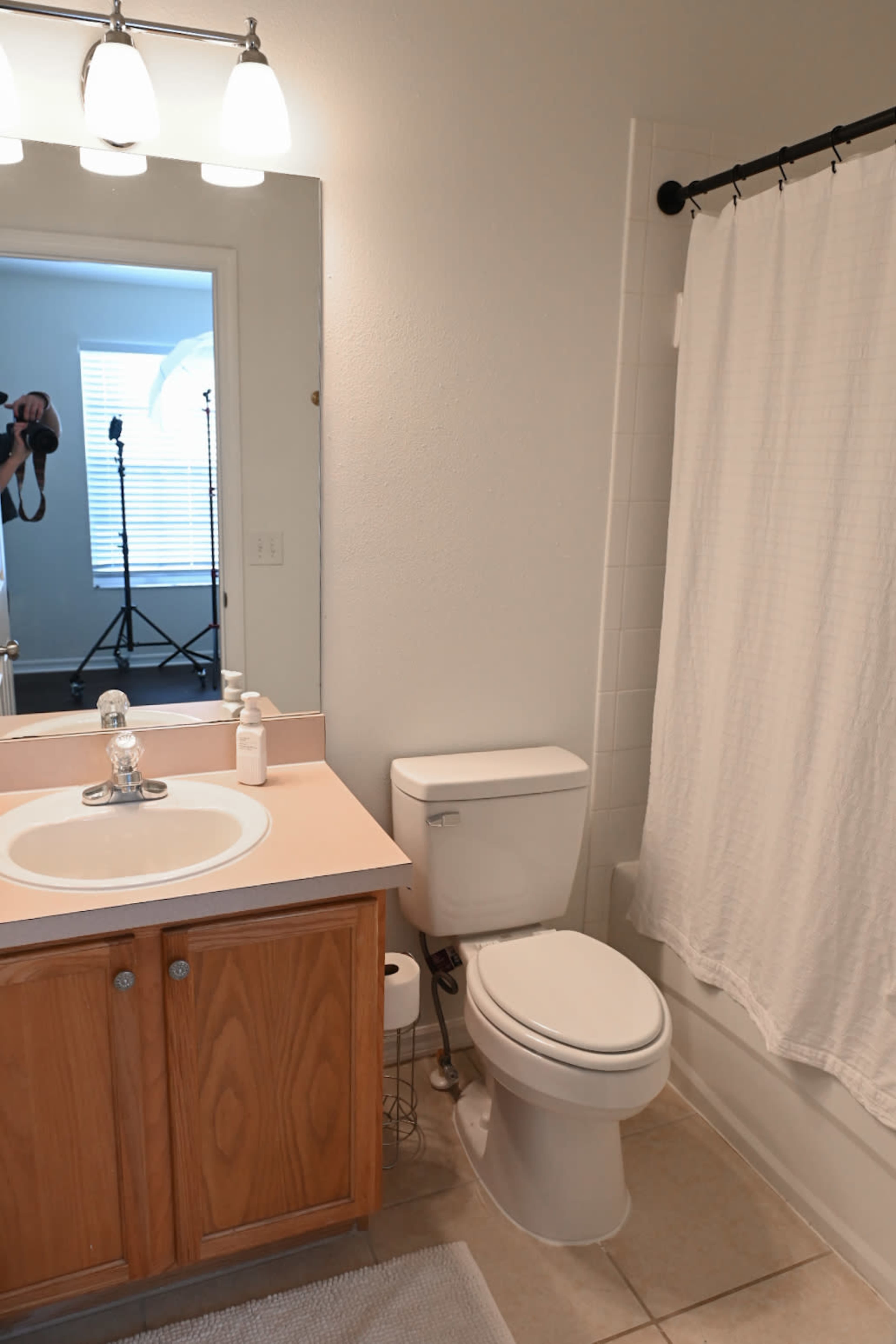 The image shows a bathroom with a white shower curtain, a sink with a mirror, and a toilet beside a wooden cabinet.