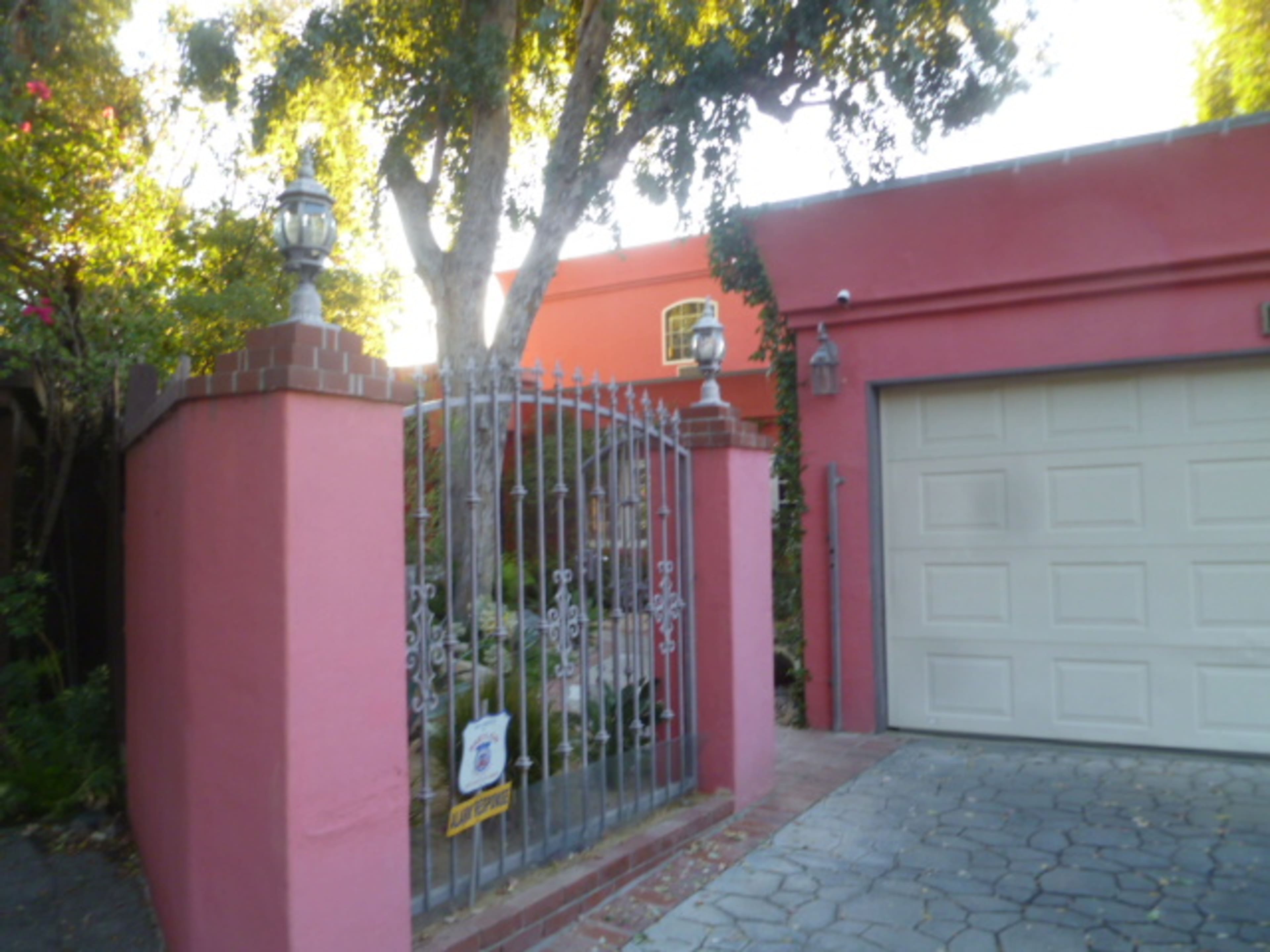 A pink stucco wall with an ornate metal gate leading to a garden and a light gray garage door beside it.
