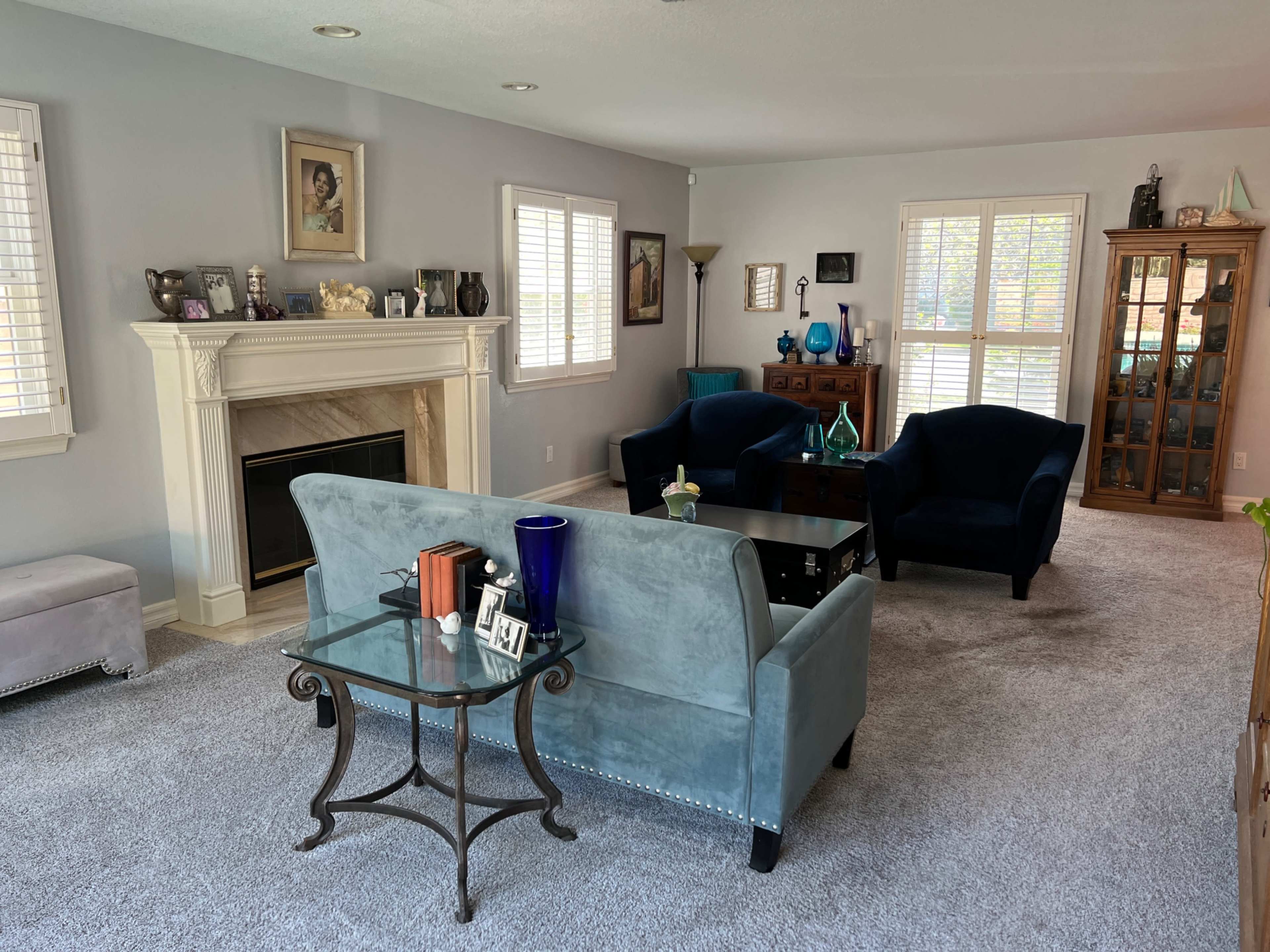 A living room with a fireplace, various pieces of furniture including a blue sofa, chairs, and a glass coffee table, all set against a neutral wall color.