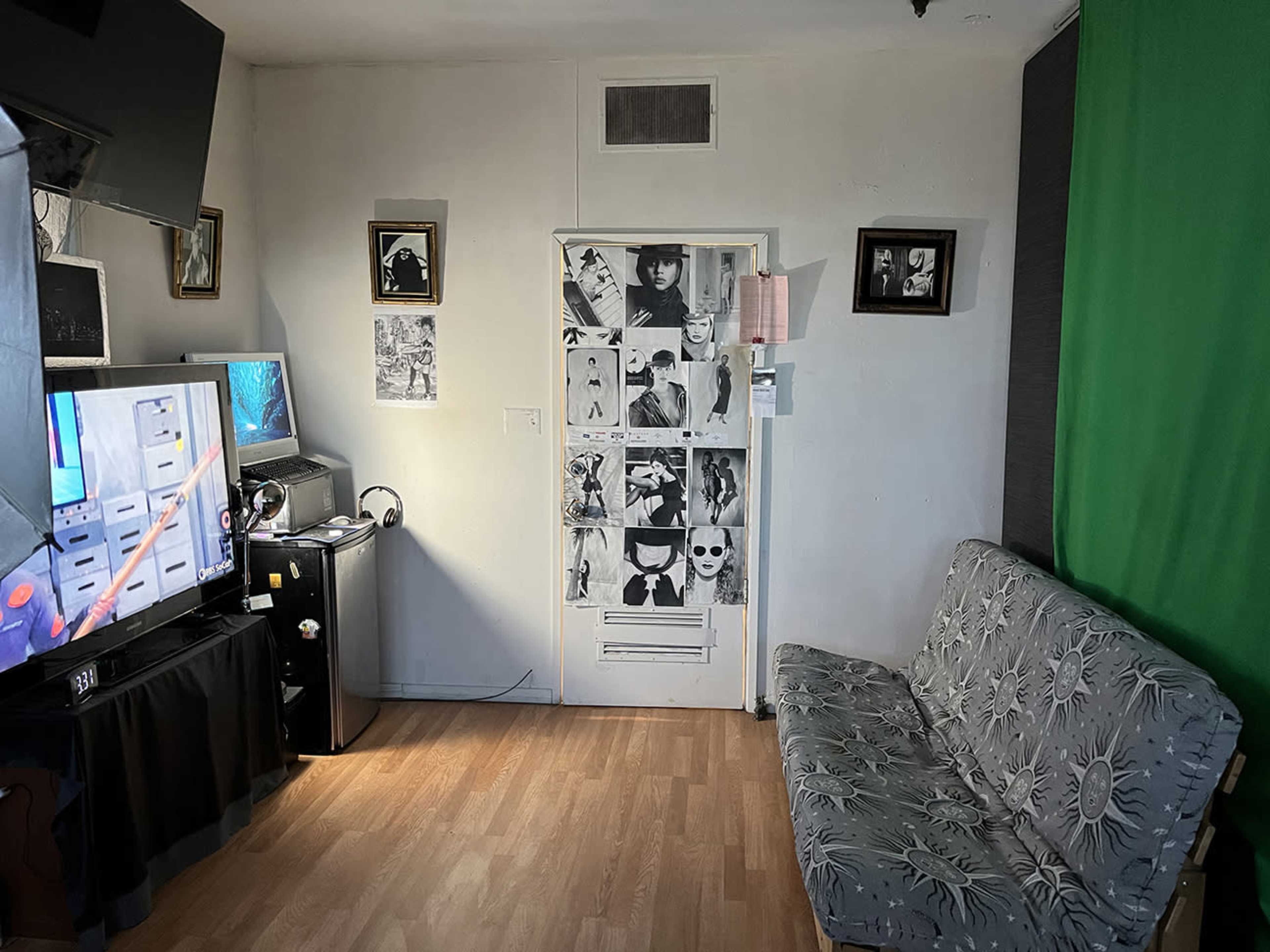 The scene features a small room with a futon, a television on a stand, a mini fridge, and a door decorated with various black and white images.