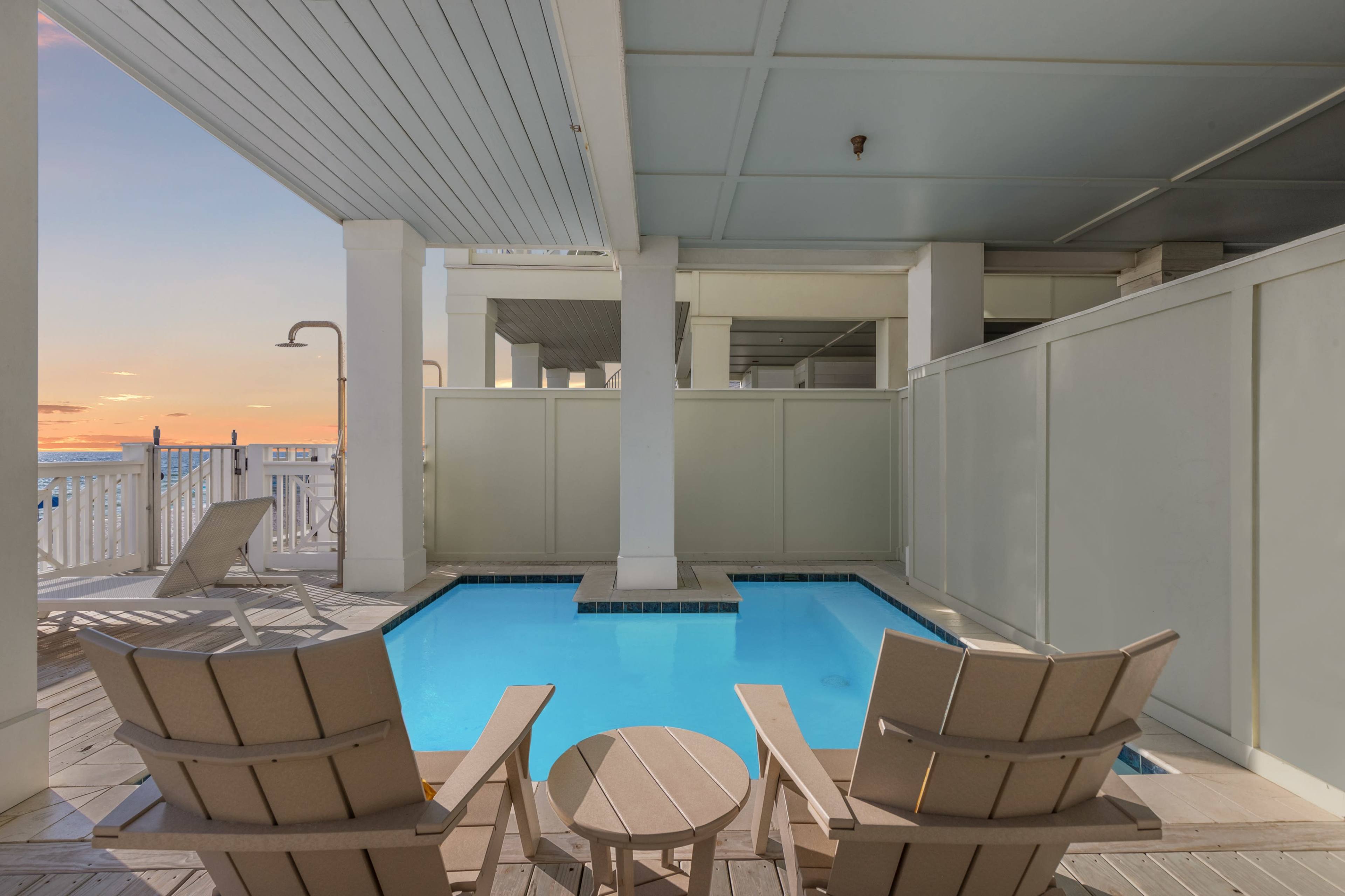 A private pool area with two wooden chairs stands next to the pool, framed by white walls and a sunset backdrop.
