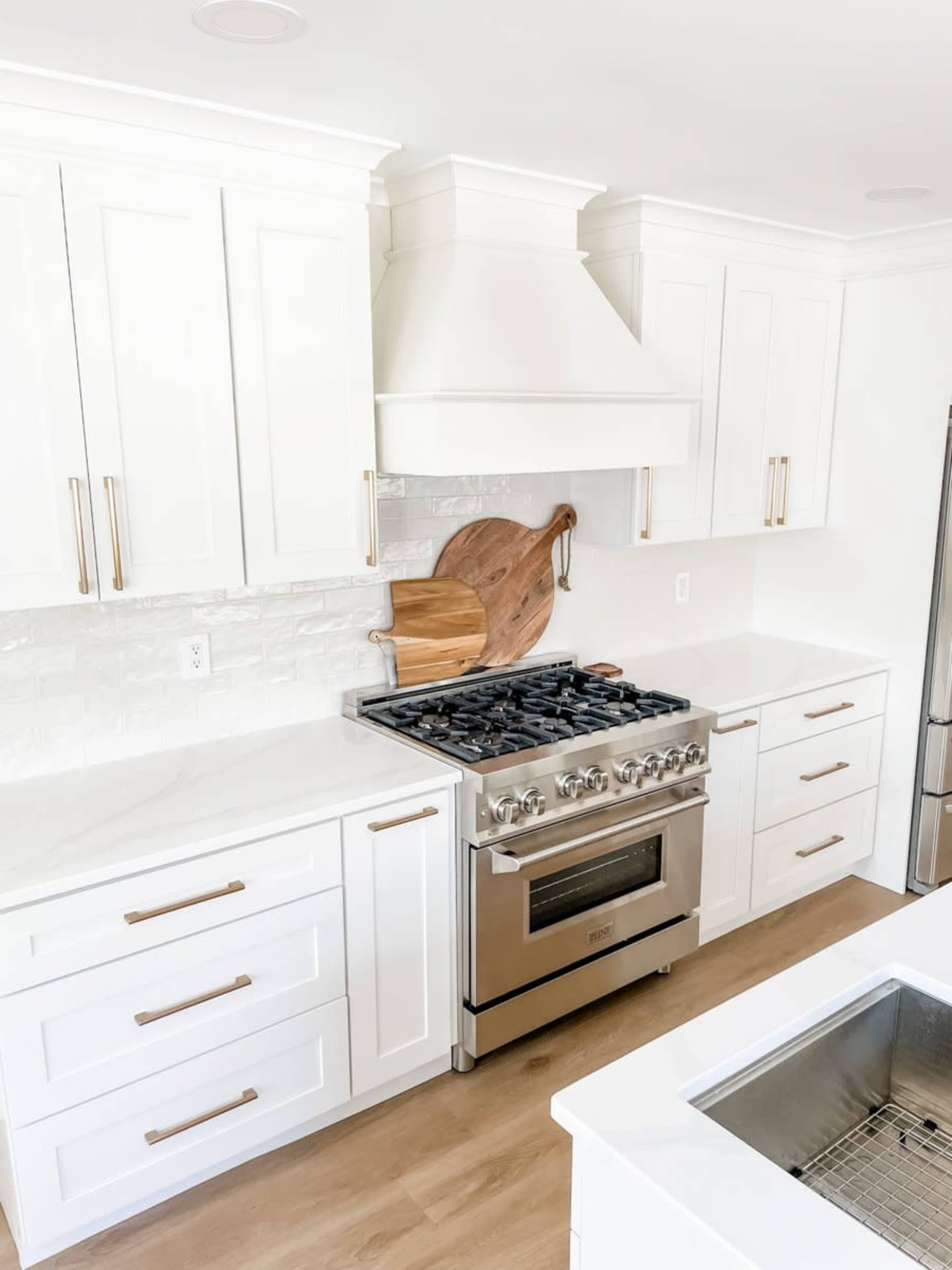 A modern kitchen featuring white cabinetry, a stainless steel stove, and a wooden cutting board leaning against the wall beneath a white hood.