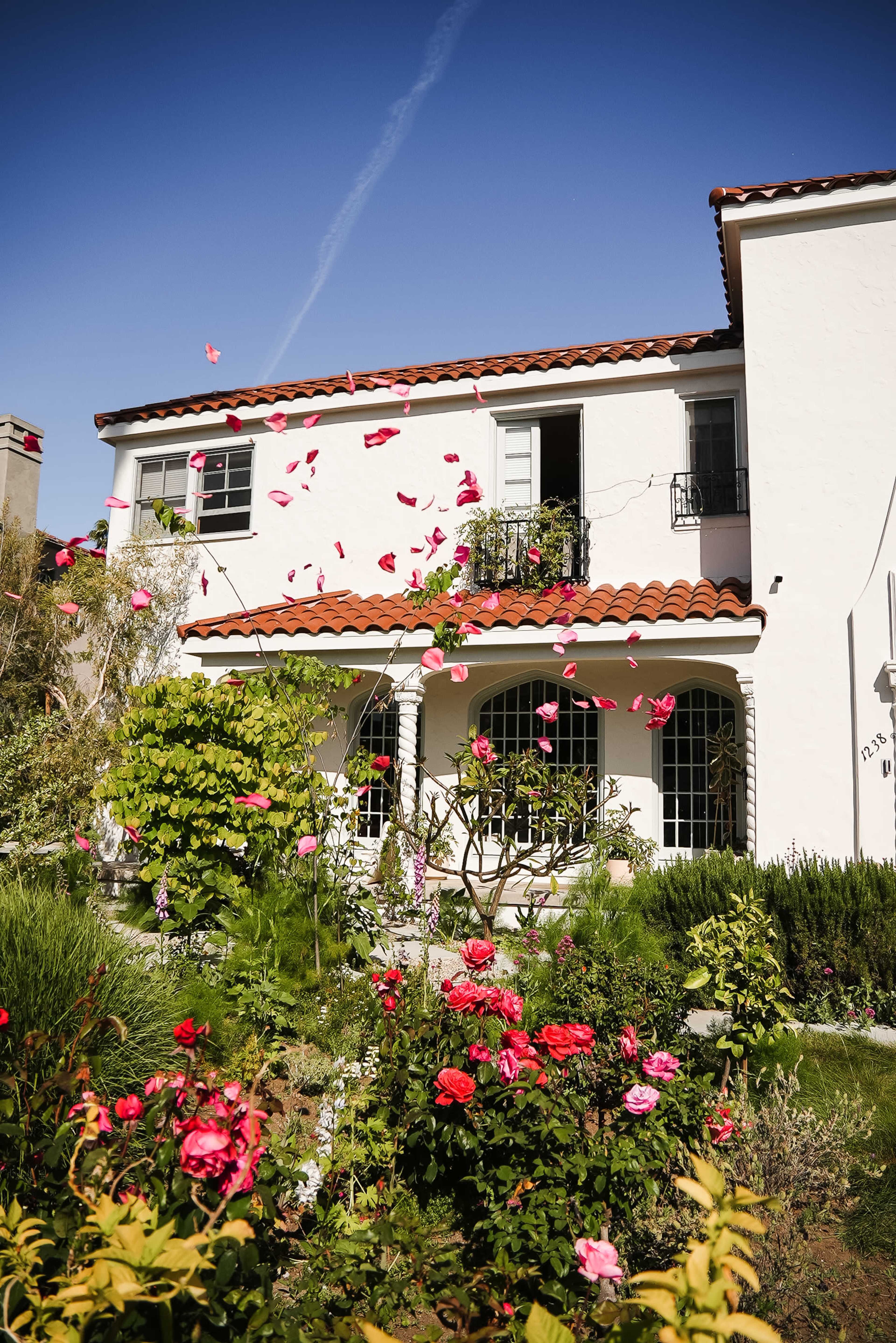 A two-story white house with a terracotta roof is surrounded by colorful rose bushes and a clear blue sky.