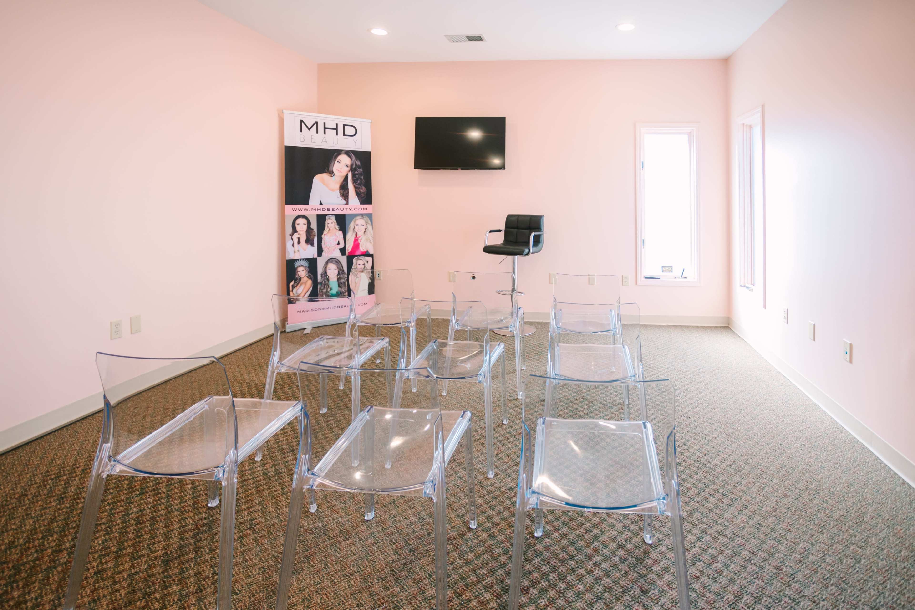A room with a light pink wall, featuring a row of clear chairs, a black stool, a television on the wall, and a banner displaying images of individuals.