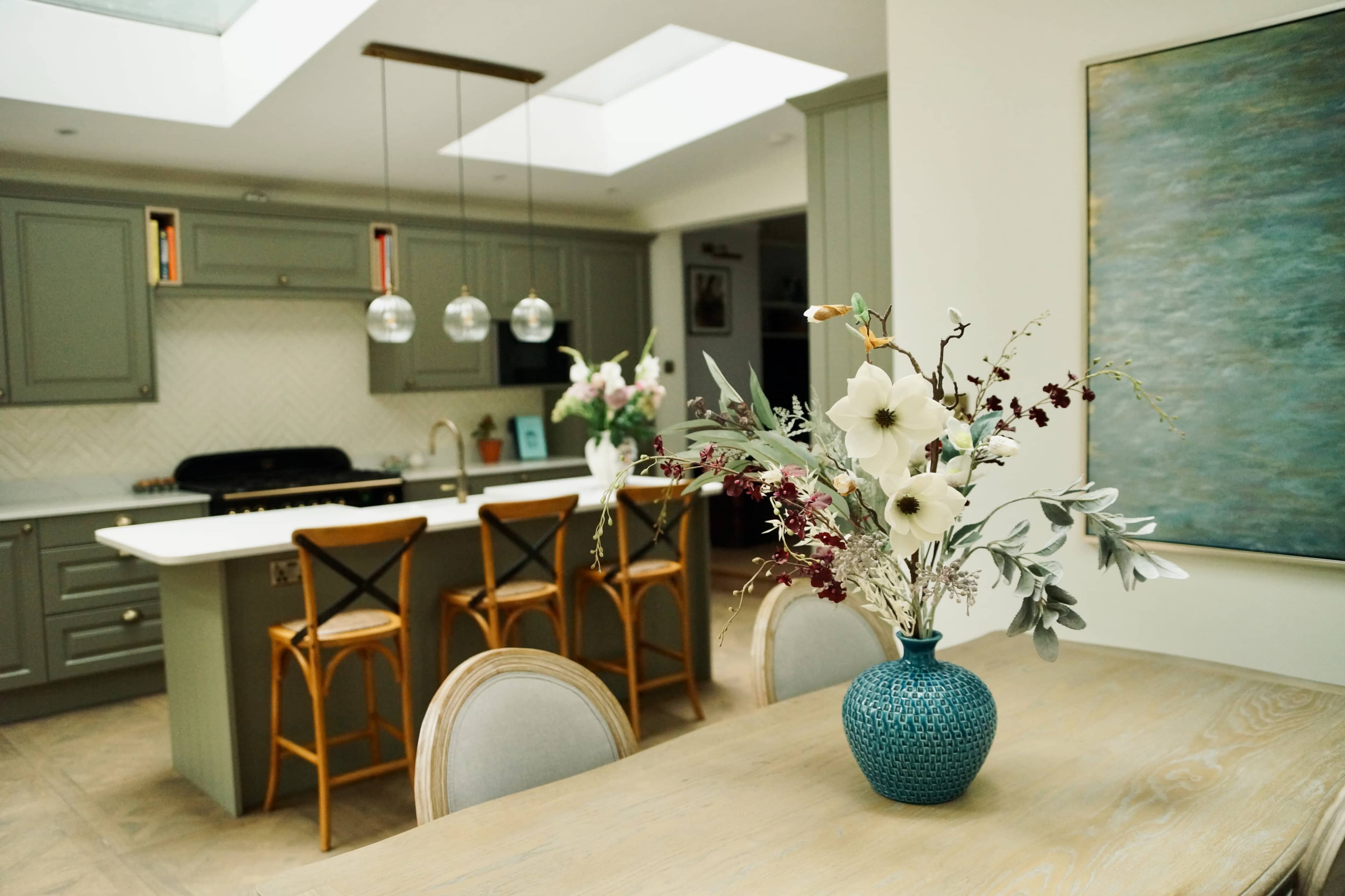 A modern kitchen with a light-filled dining area, featuring a vase of flowers on a wooden table and bar stools at the counter.