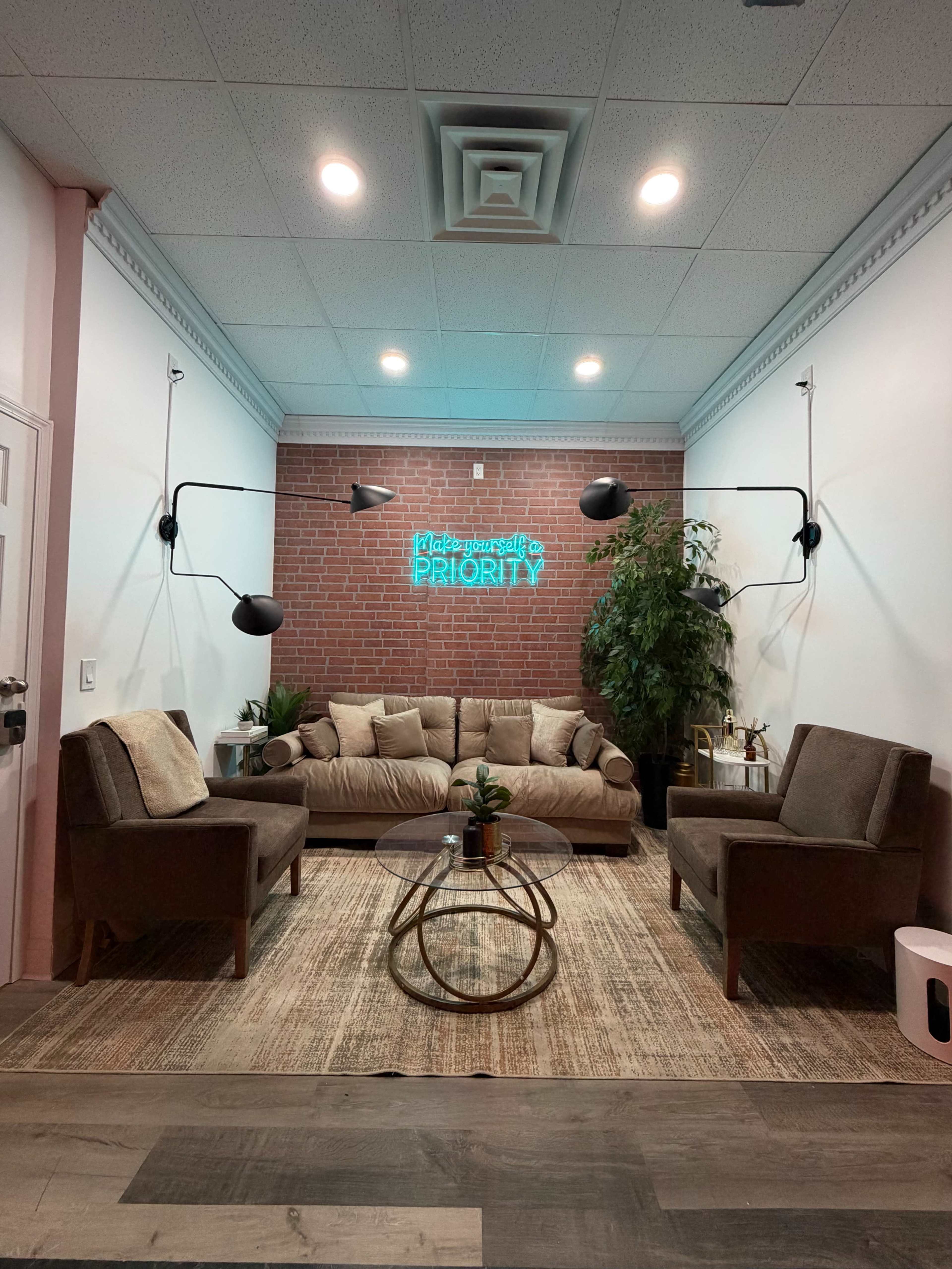 The image shows a cozy lounge area with a brown sofa, two armchairs, a round coffee table, and greenery, highlighted by a neon sign on a brick wall.