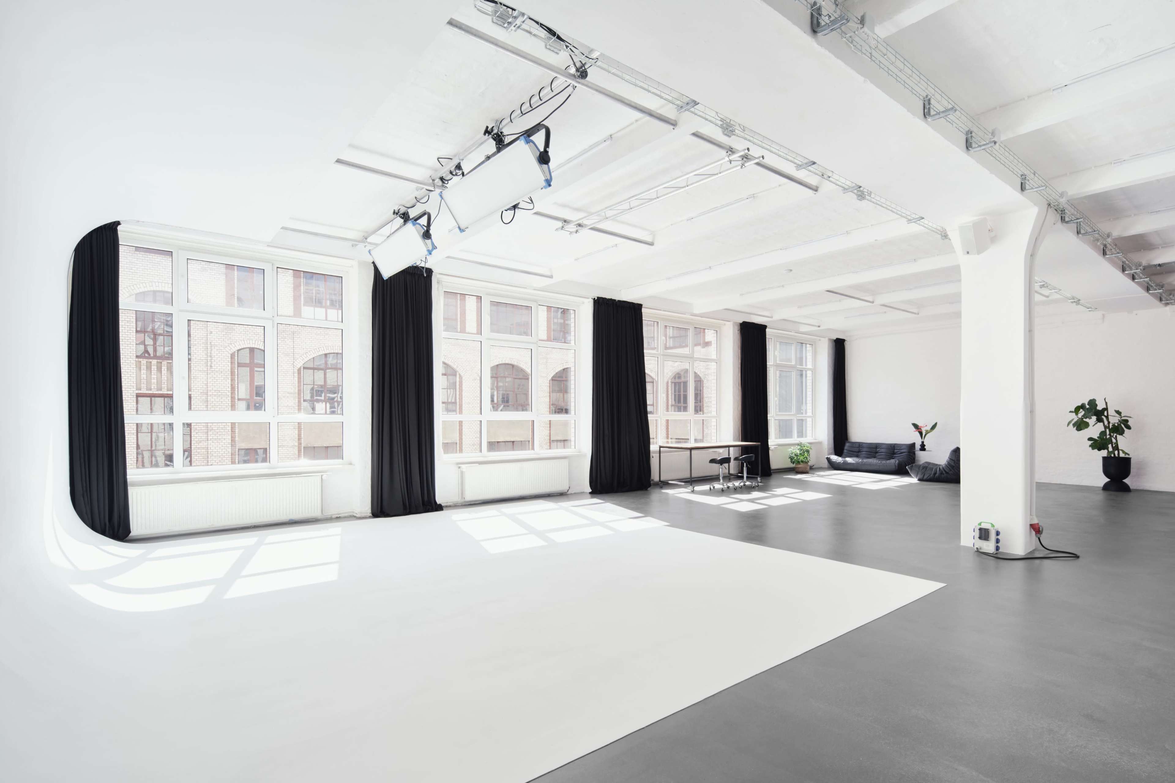 The image shows a bright, spacious studio with large windows, white walls, and a minimalistic interior featuring a black couch, a potted plant, and a light backdrop area.